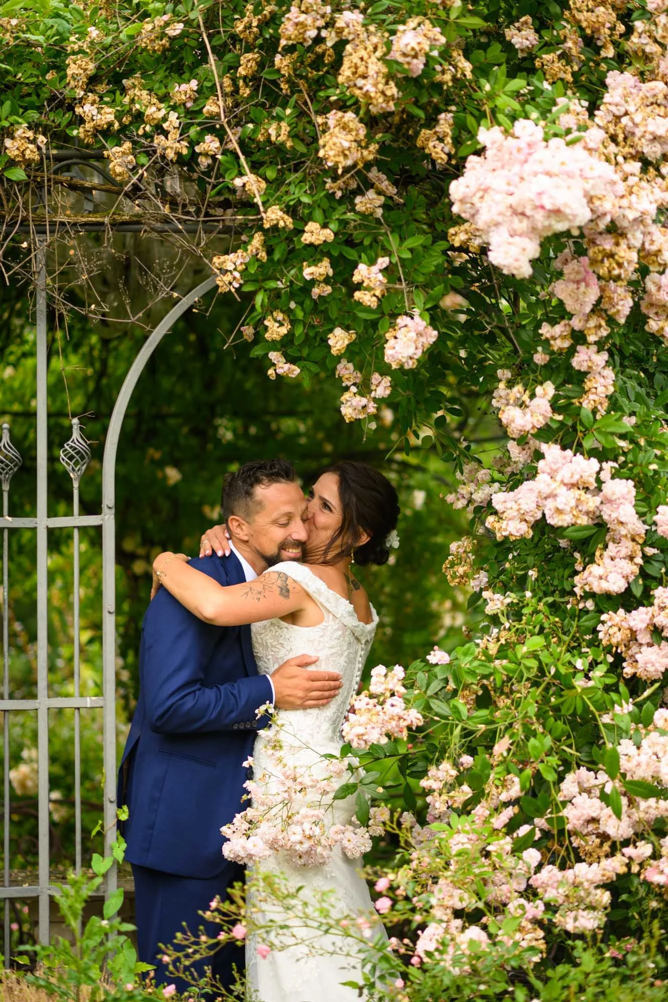 Un couple de mariés s'embrassant dans un jardin entouré de rosiers en fleurs, sous une arche en métal.