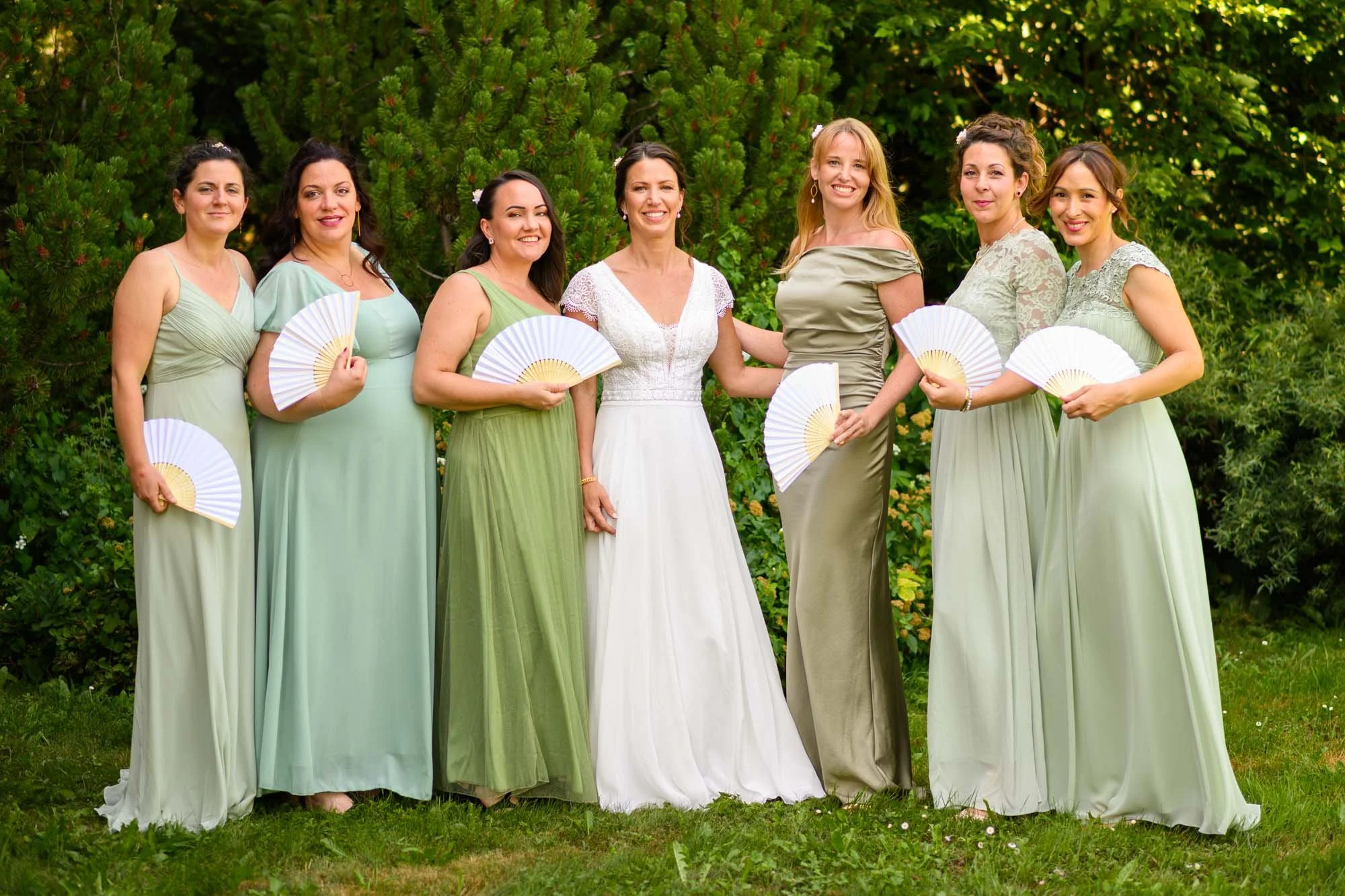 Groupe de femmes en robes vertes et blanche, posant en plein air avec des éventails, devant des arbustes et des arbres.