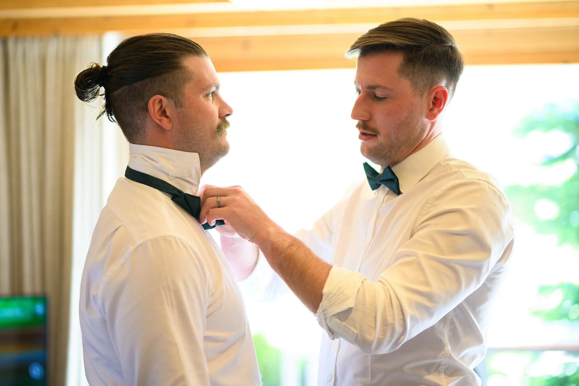 Deux hommes en chemises blanches et nœuds papillons noirs, l'un face à l'autre, dans une pièce lumineuse, l'un ajustant la cravate de l'autre.