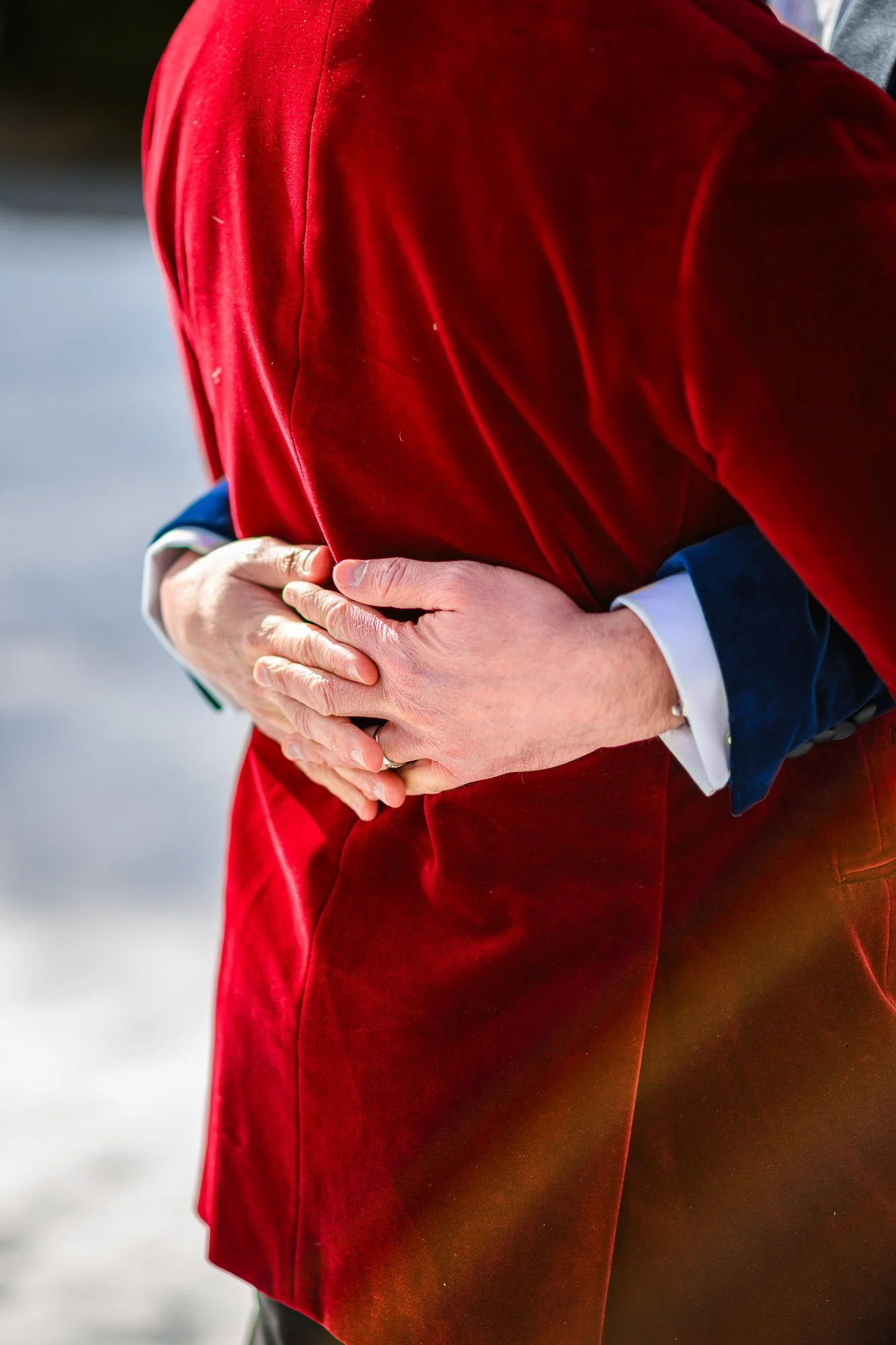 Deux personnes en costume, l'une en manteau rouge et l'autre en costume bleu, se tiennent au centre de l'image, tenant la main de l'autre au niveau du ventre. La scène se déroule probablement en extérieur sur fond lumineux.