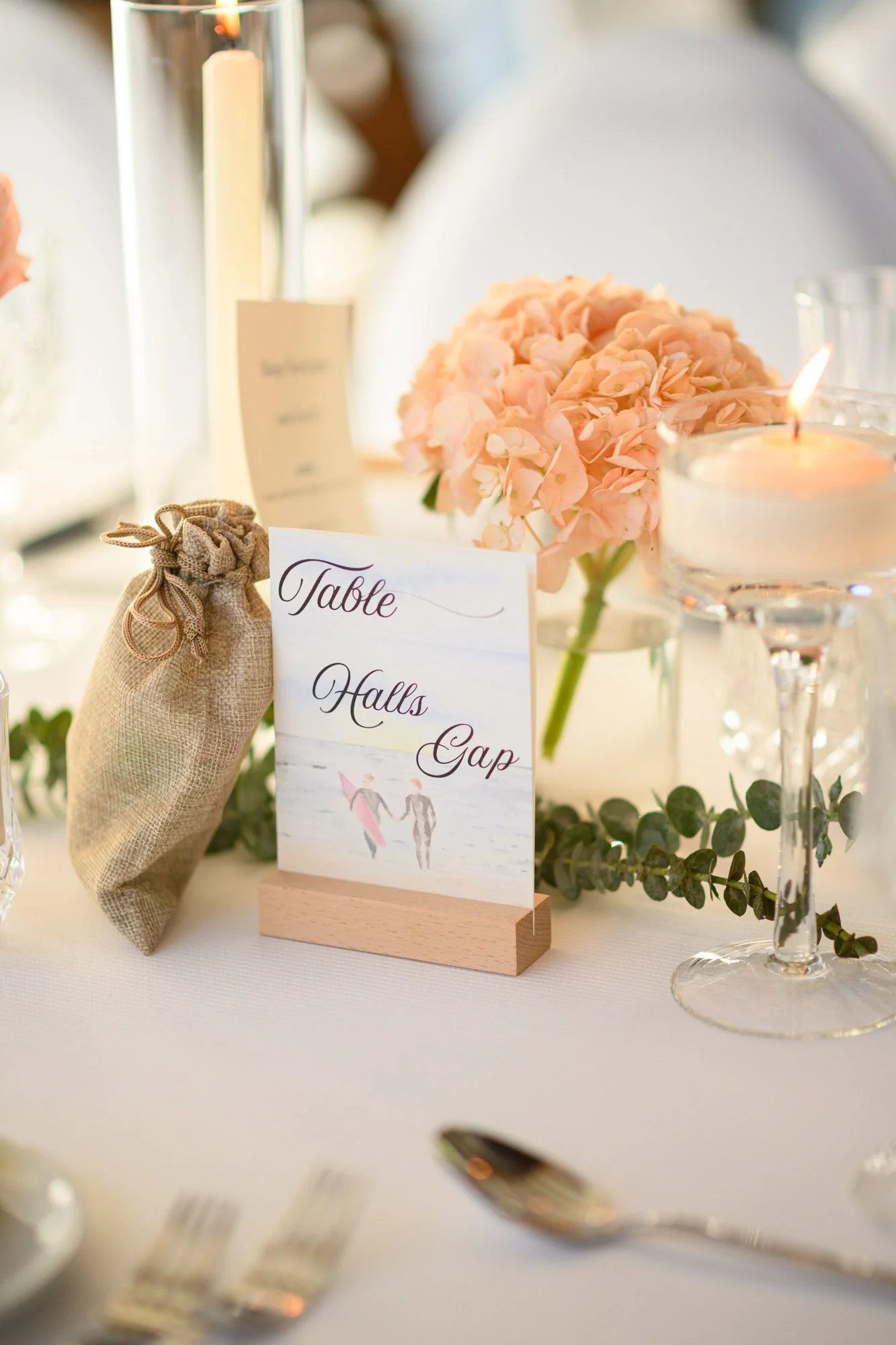 Table de mariage décorée avec une composition florale de hortensias rose pâle, des bougies, un sachet en toile, et une carte indiquant les tables, séparées par un espace en forme de vide.