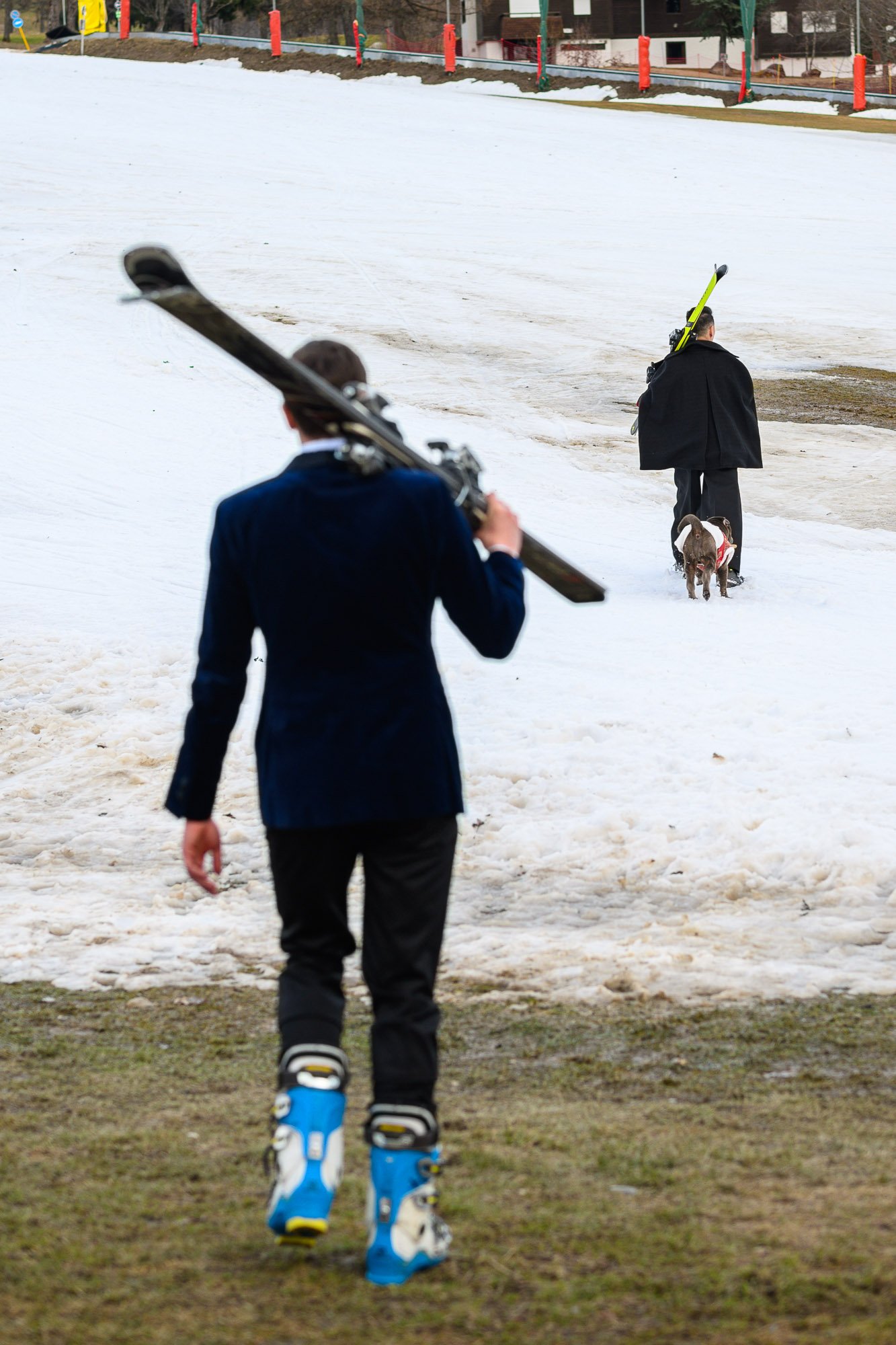 Deux hommes, l'un portant des skis et l'autre avec un chien, marchent sur une pente enneigée, le premier vu de dos avec des bottes de ski bleues, le second plus éloigné avec un manteau noir, en direction d'une maison en arrière-plan.