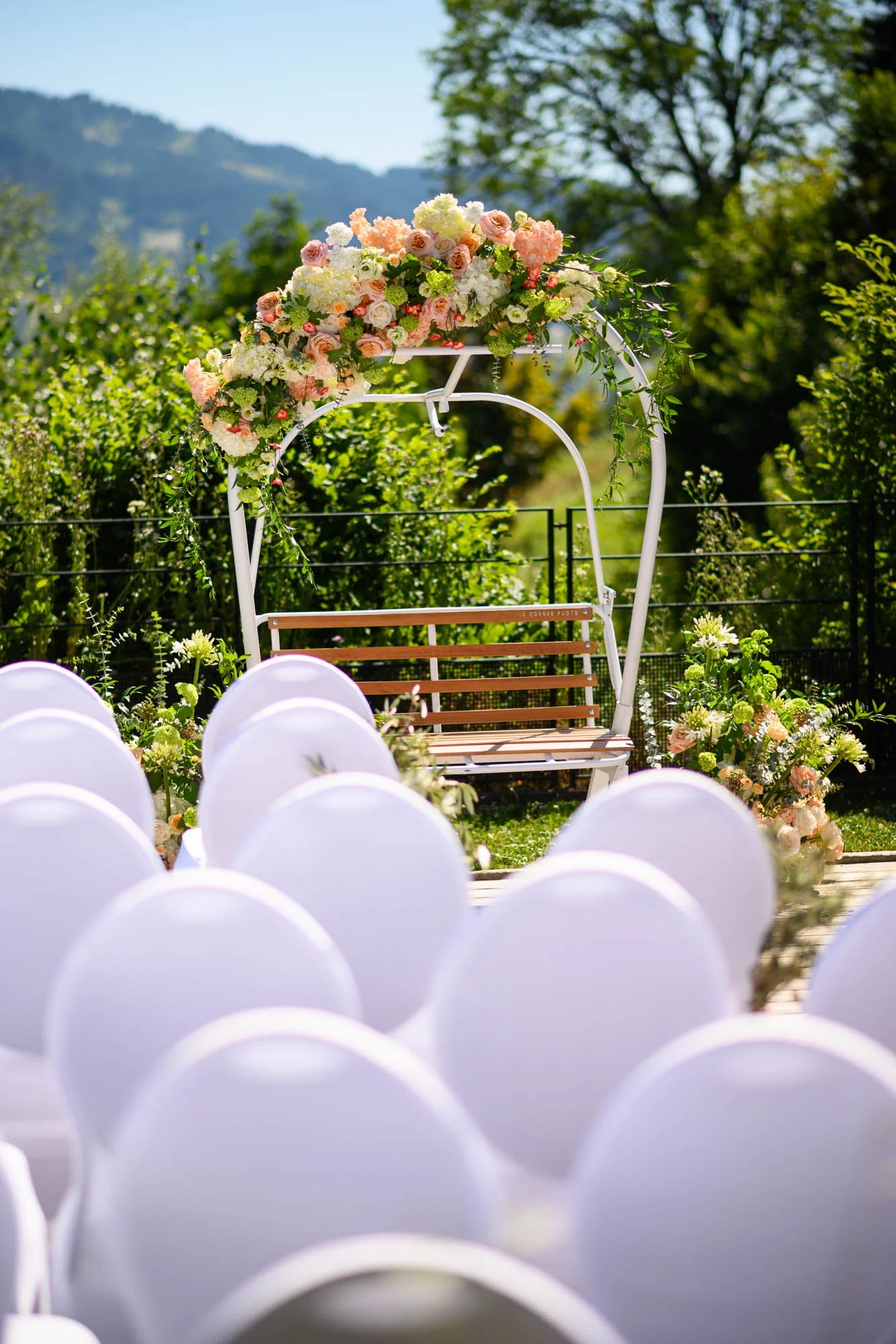 Décor de mariage en plein air avec une balançoire ornée de fleurs pastel, entourée de chaises blanches et de bouquets de fleurs. Paysage verdoyant en arrière-plan.