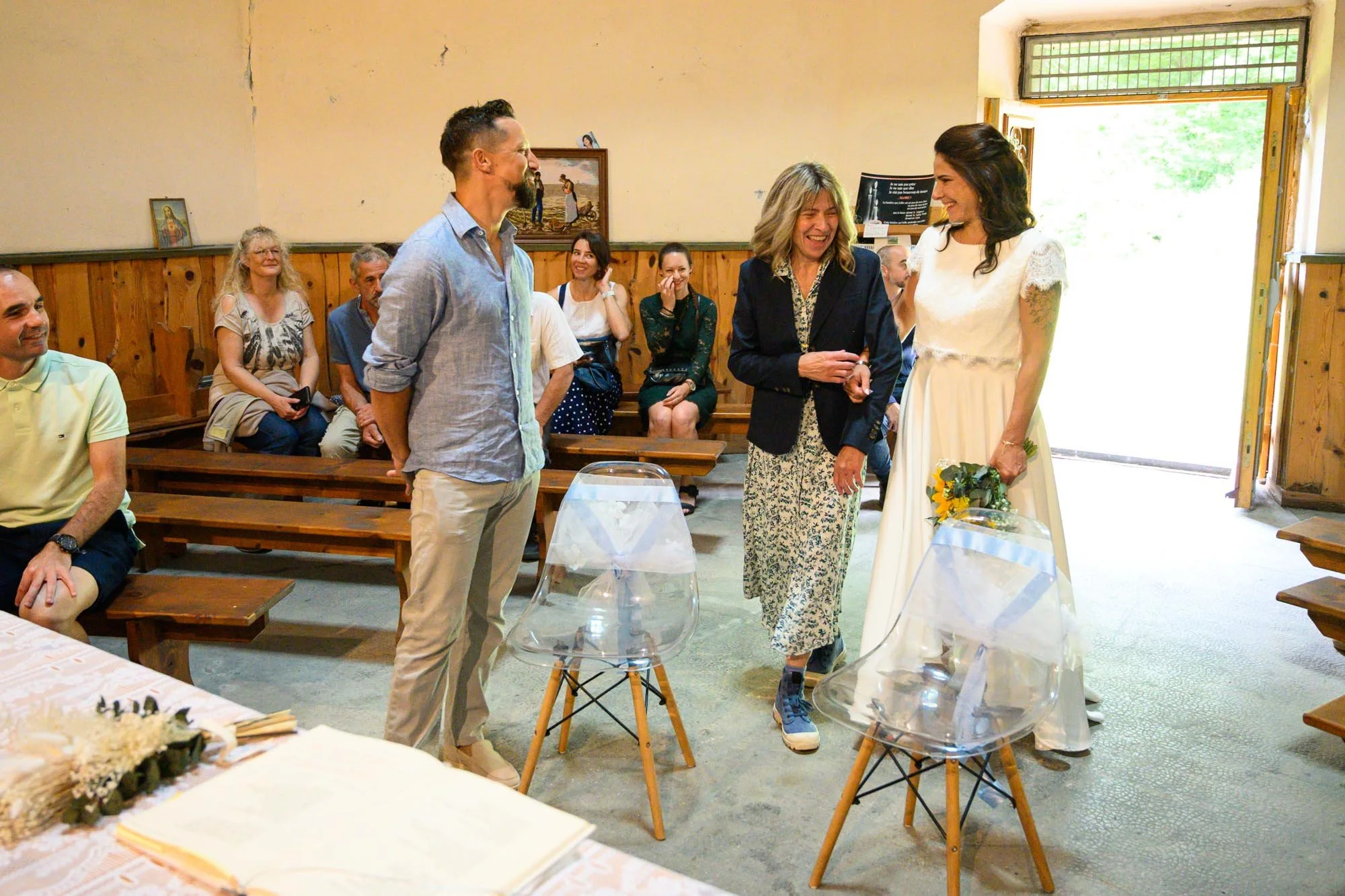 Un groupe de personnes souriantes partage un moment joyeux dans une pièce, probablement lors d'une cérémonie ou d'une célébration. Au centre, une femme en robe blanche tient un bouquet de fleurs, entourée de personnes qui semblent lui adresser des co