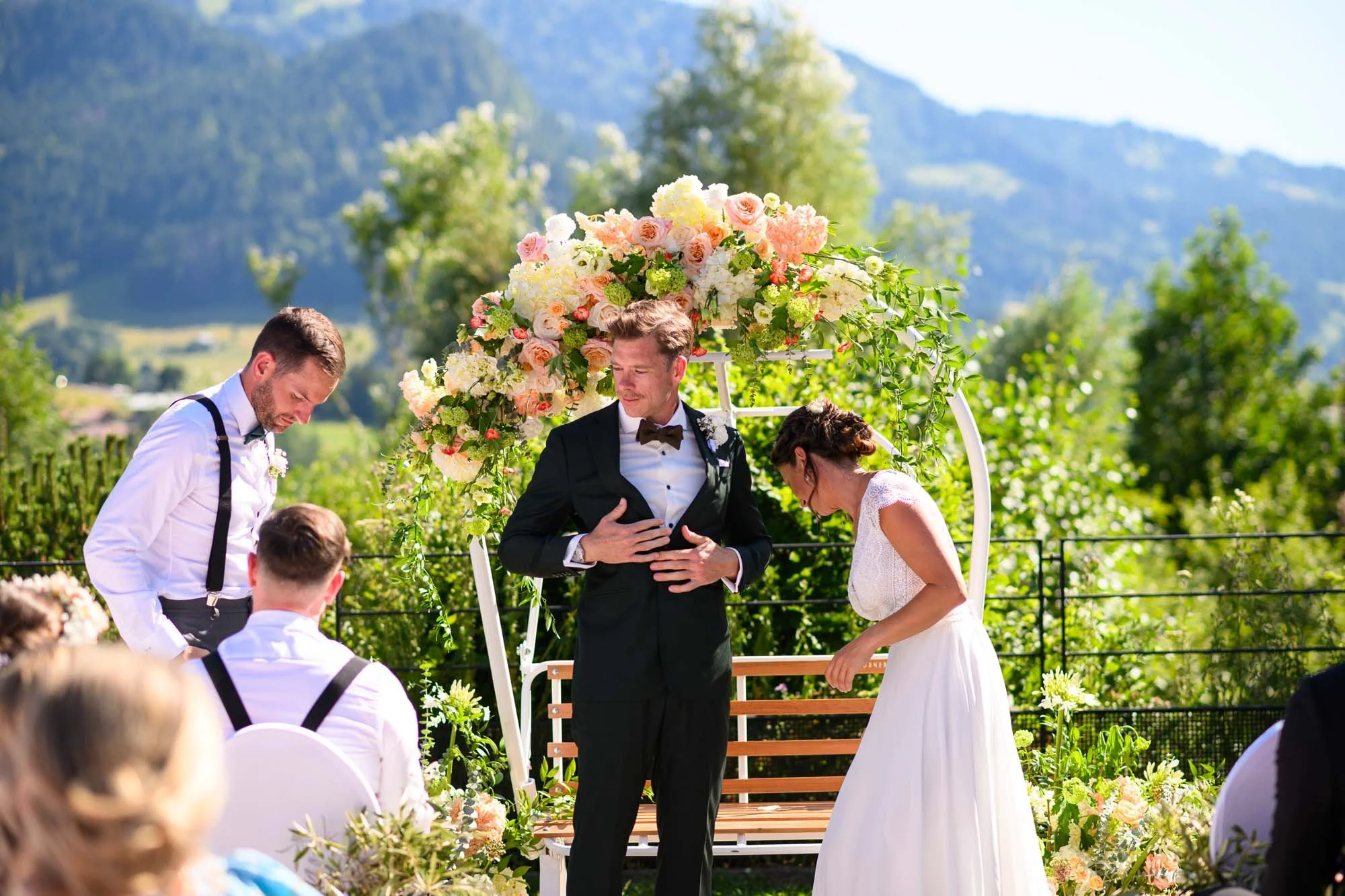 Un couple marié lors d'une cérémonie en plein air, entourés de fleurs et de végétation derrière une arche décorée de fleurs, avec des invités assis à proximité.