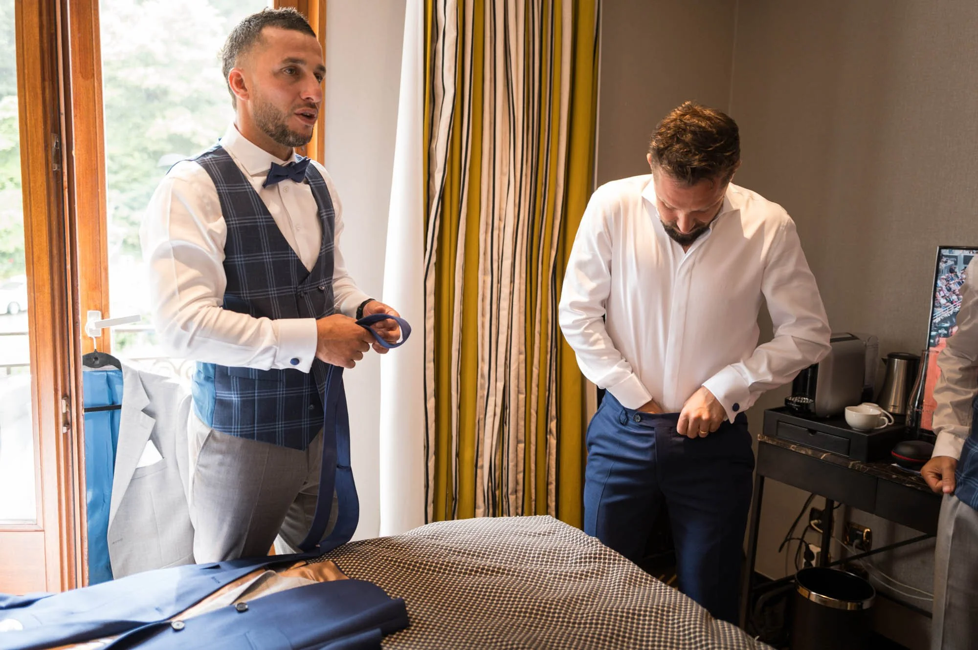 Deux hommes en tenue de mariage se préparent dans une chambre, l'un met un pantalon et l'autre tient une cravate, avec une valise ouverte sur un lit et un coin café en arrière-plan.