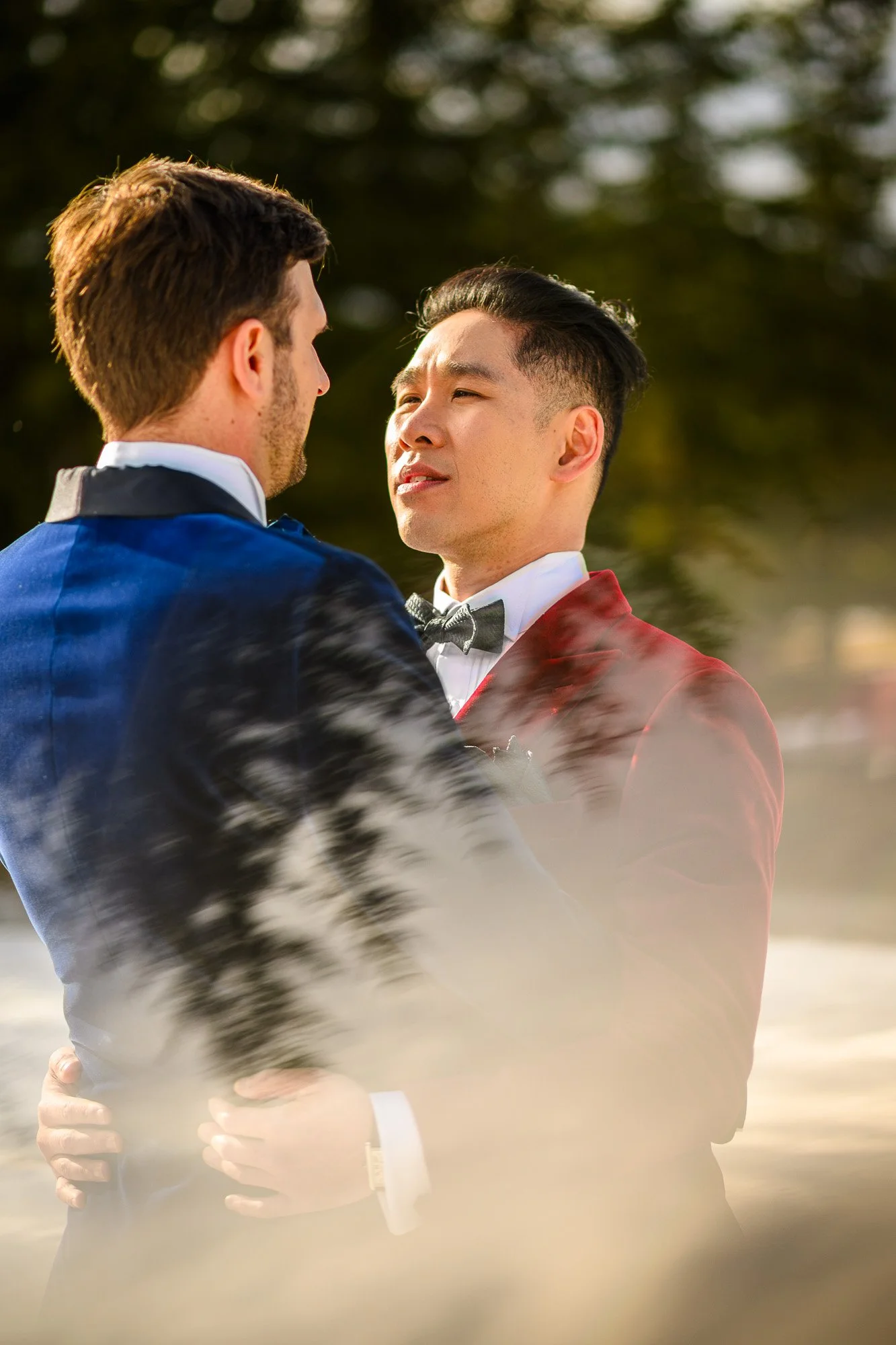 Deux hommes habillés en costumes de soirée, dansant ensemble à l'extérieur, avec un fond flou d'arbres et un ciel ensoleillé.