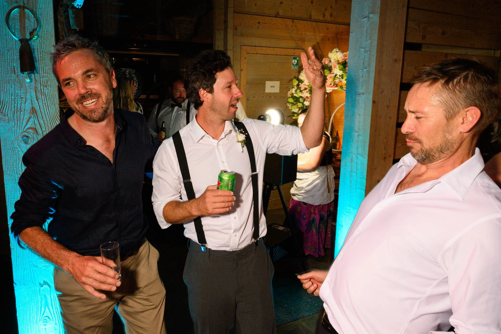 Trois hommes lors d'une célébration, deux d'entre eux sont en train de discuter, l'un tient une canette, l'autre un verre. Un arrière-plan avec des fleurs et un décor en bois