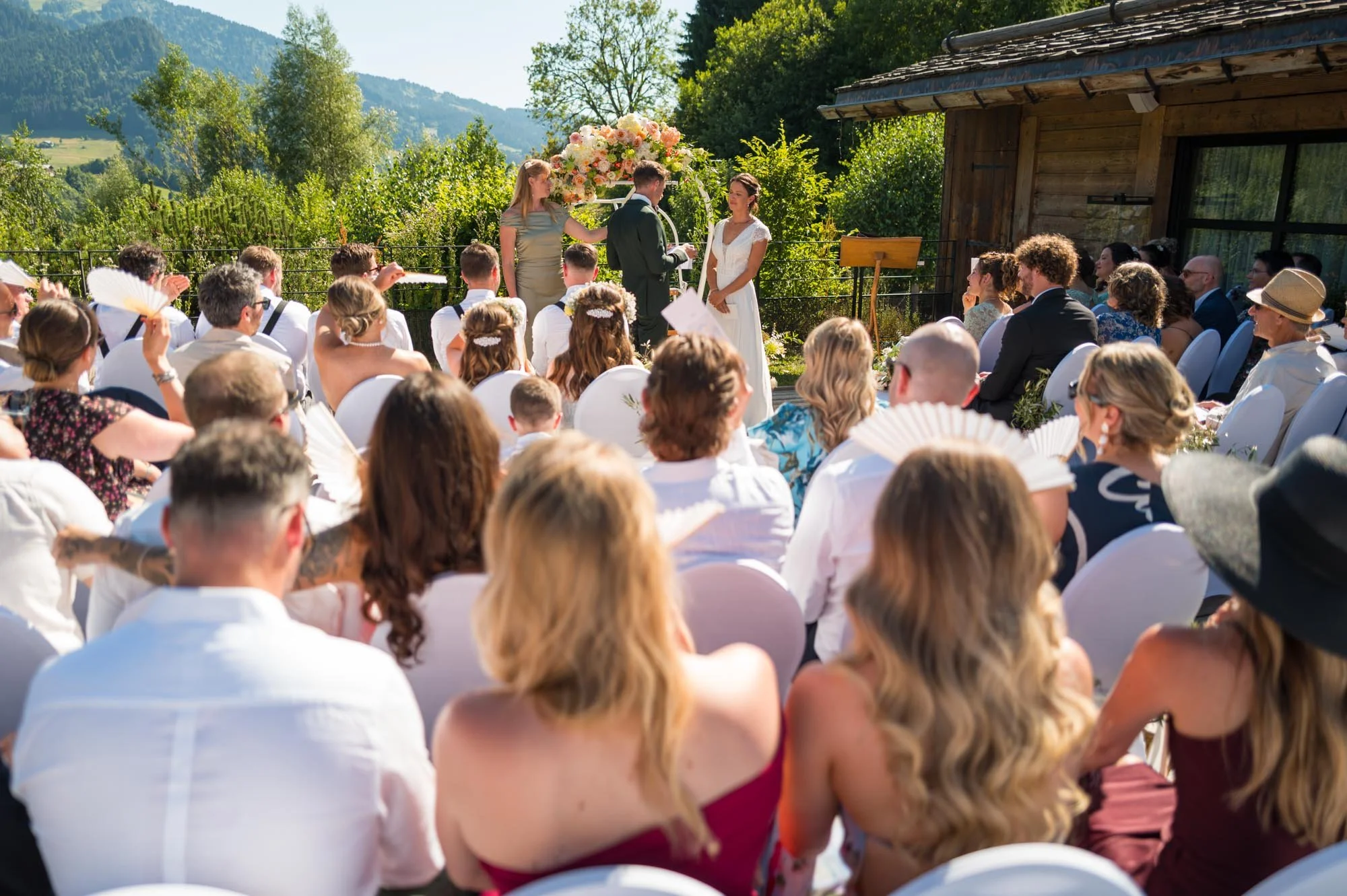 Cérémonie de mariage en plein air avec un couple marié, officiant, et invités assis en rangée, dans un décor naturel avec montagnes et végétation, sous un ciel ensoleillé.