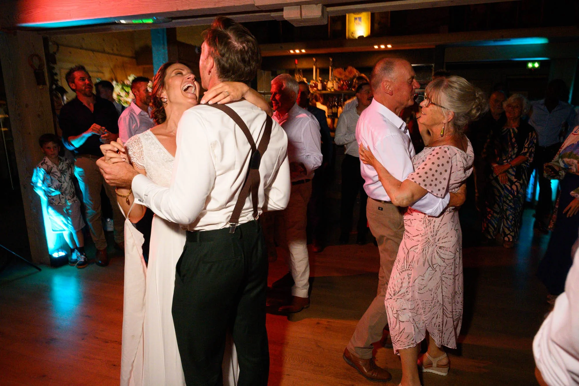 Un groupe de personnes dansant et souriant lors d'une fête ou d'un mariage, avec plusieurs couples en train de danser en arrière-plan, ambiance festive et chaleureuse.