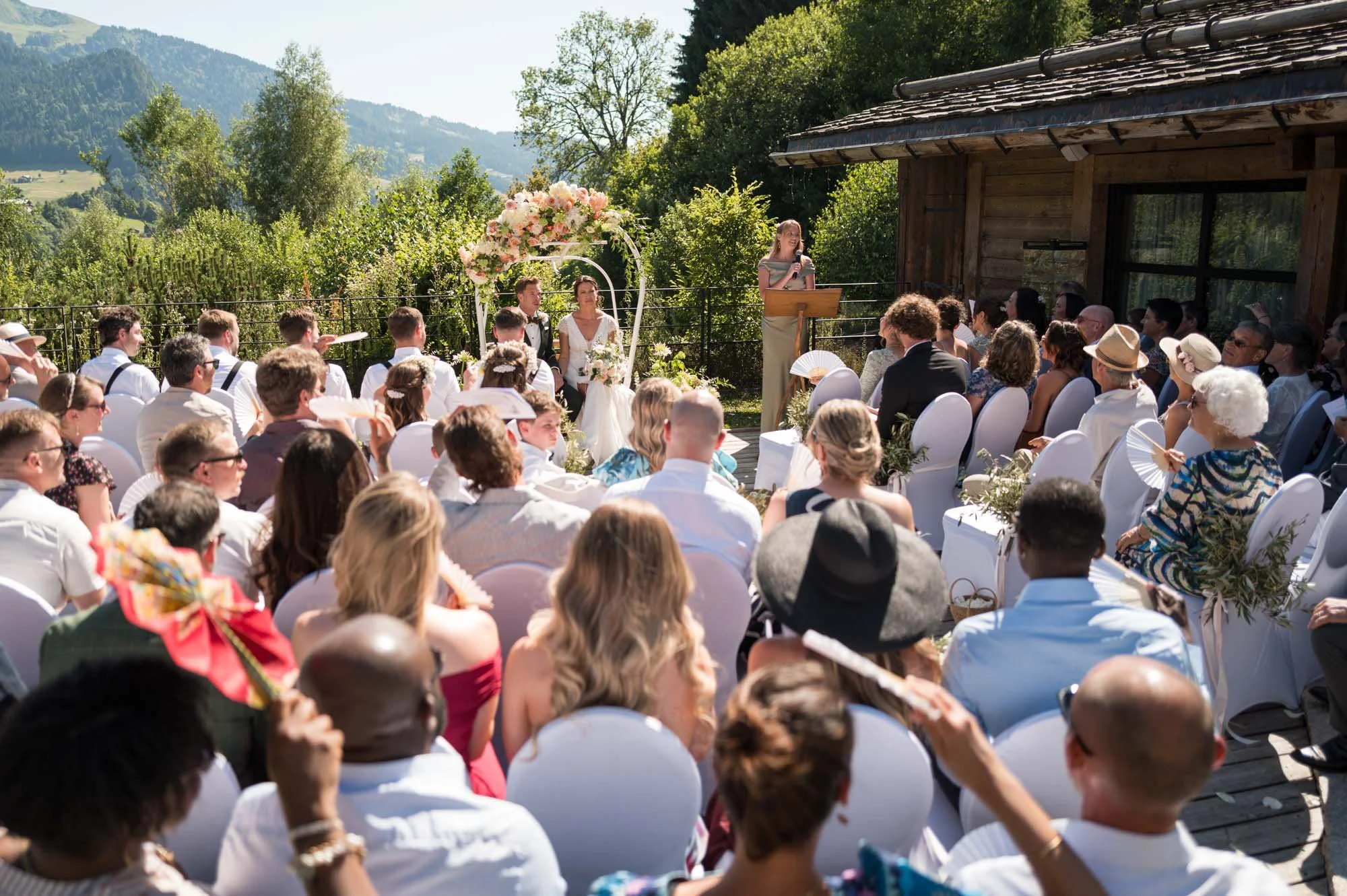 Cérémonie de mariage en plein air avec un groupe d'invités assis, un couple de mariés assis sous une arche de fleurs, une femme parlant au micro, fond de montagnes et de verdure.