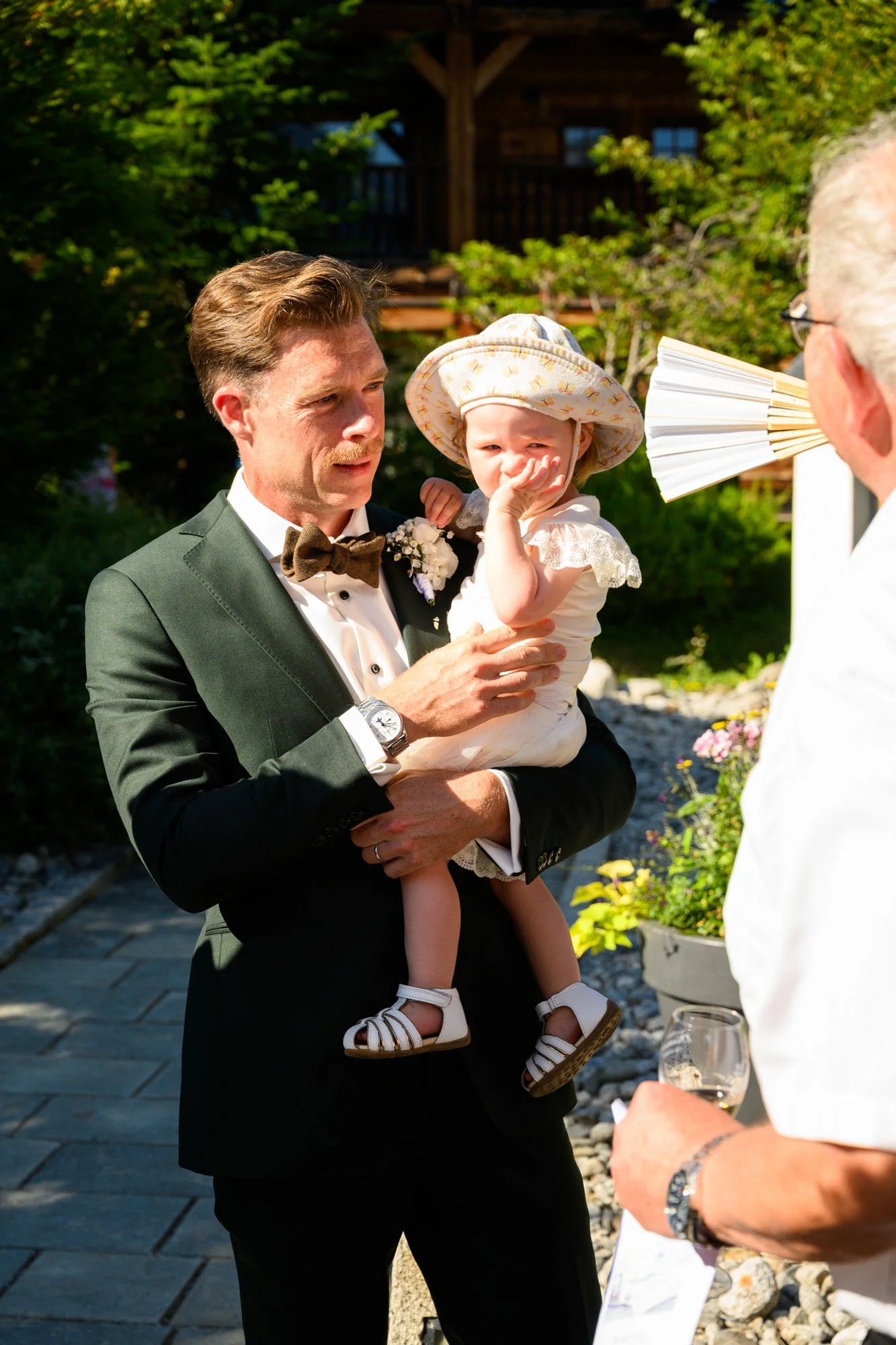 Homme en smoking tenant une jeune fille, lors d'une célébration en plein air, avec un arrière-plan de nature verte.