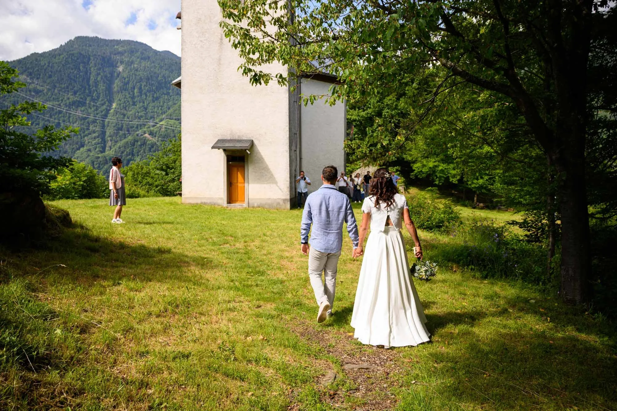 Un couple en mariage marche dans un espace vert, à proximité d'une église en montagne, avec des invités en arrière-plan