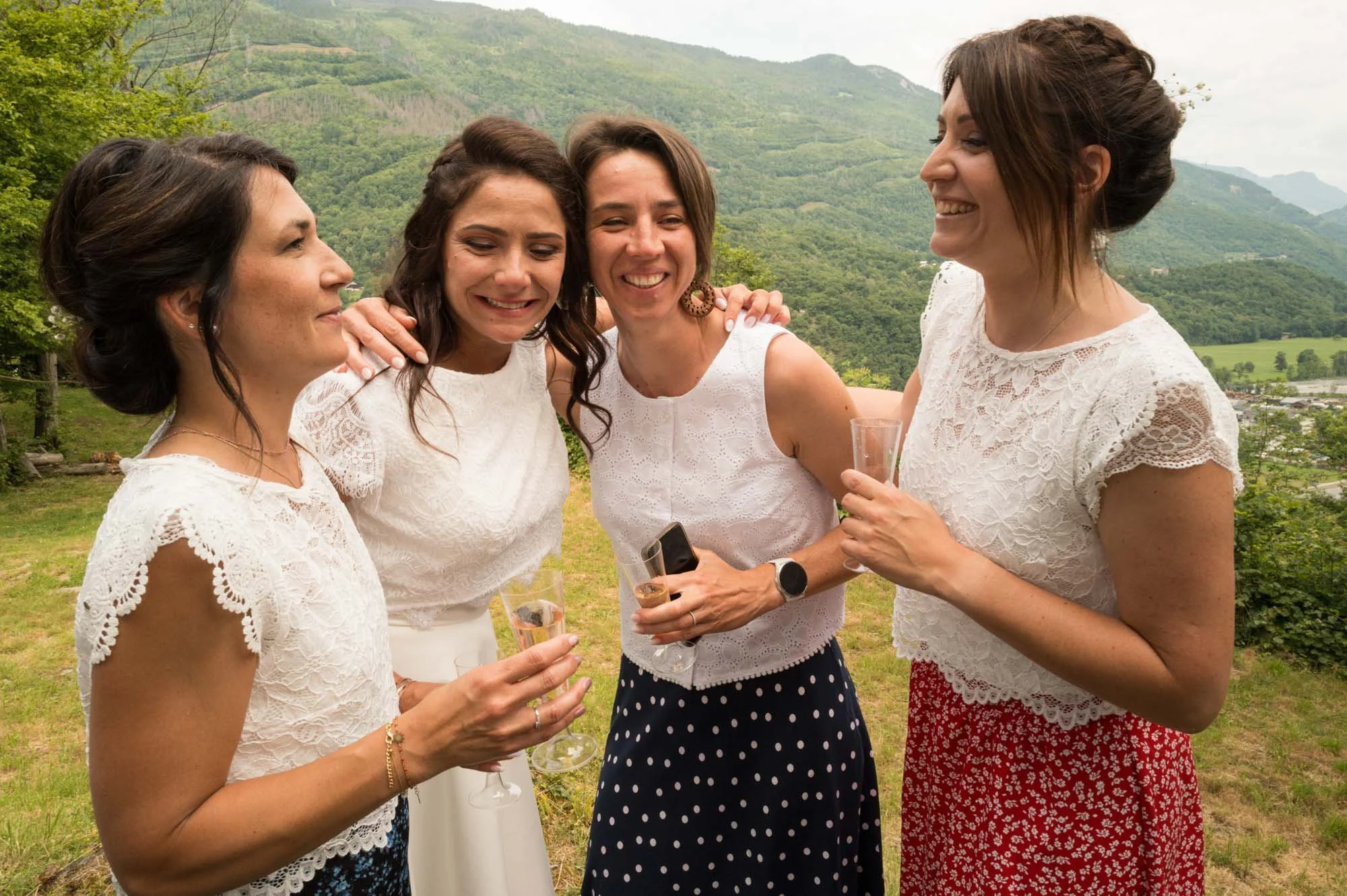 Quatre femmes souriantes en tenues blanches et colorées, tenant des flûtes à champagne, lors d'un rassemblement en plein air avec un paysage de montagnes en arrière-plan.