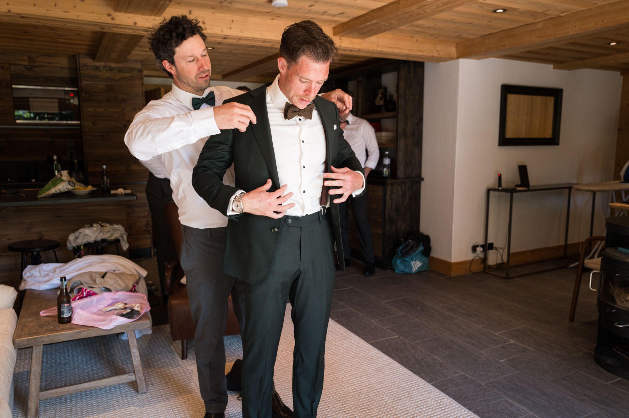 Deux hommes en costume se préparent pour une cérémonie, un ajustant le blazer de l'autre dans une pièce décorée en bois.