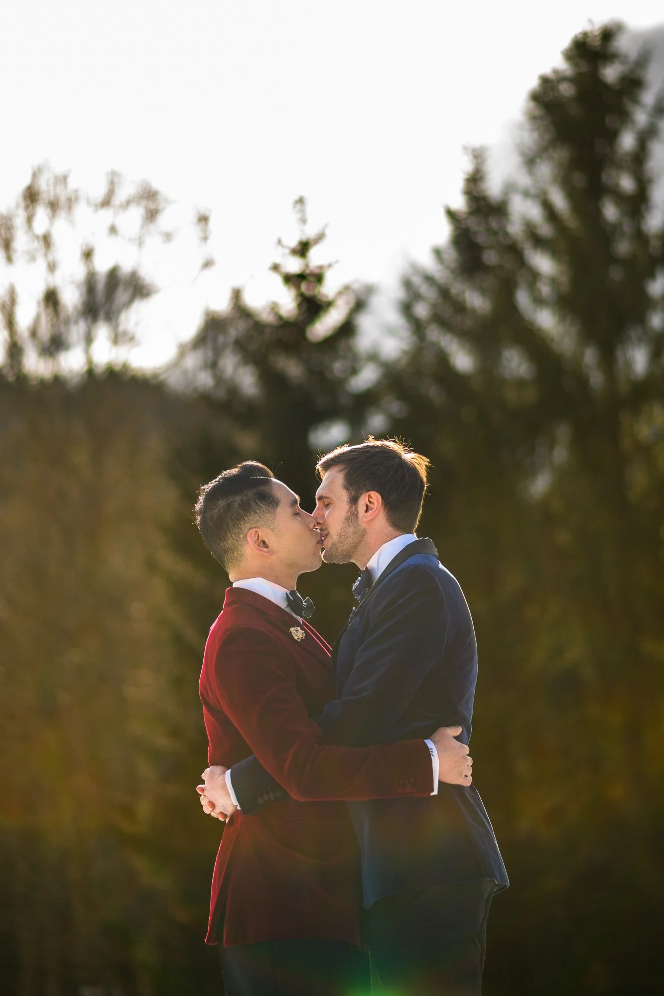 Deux hommes s'embrassent dans un parc, portant des costumes formels, avec des arbres en arrière-plan et la lumière du soleil derrière eux.