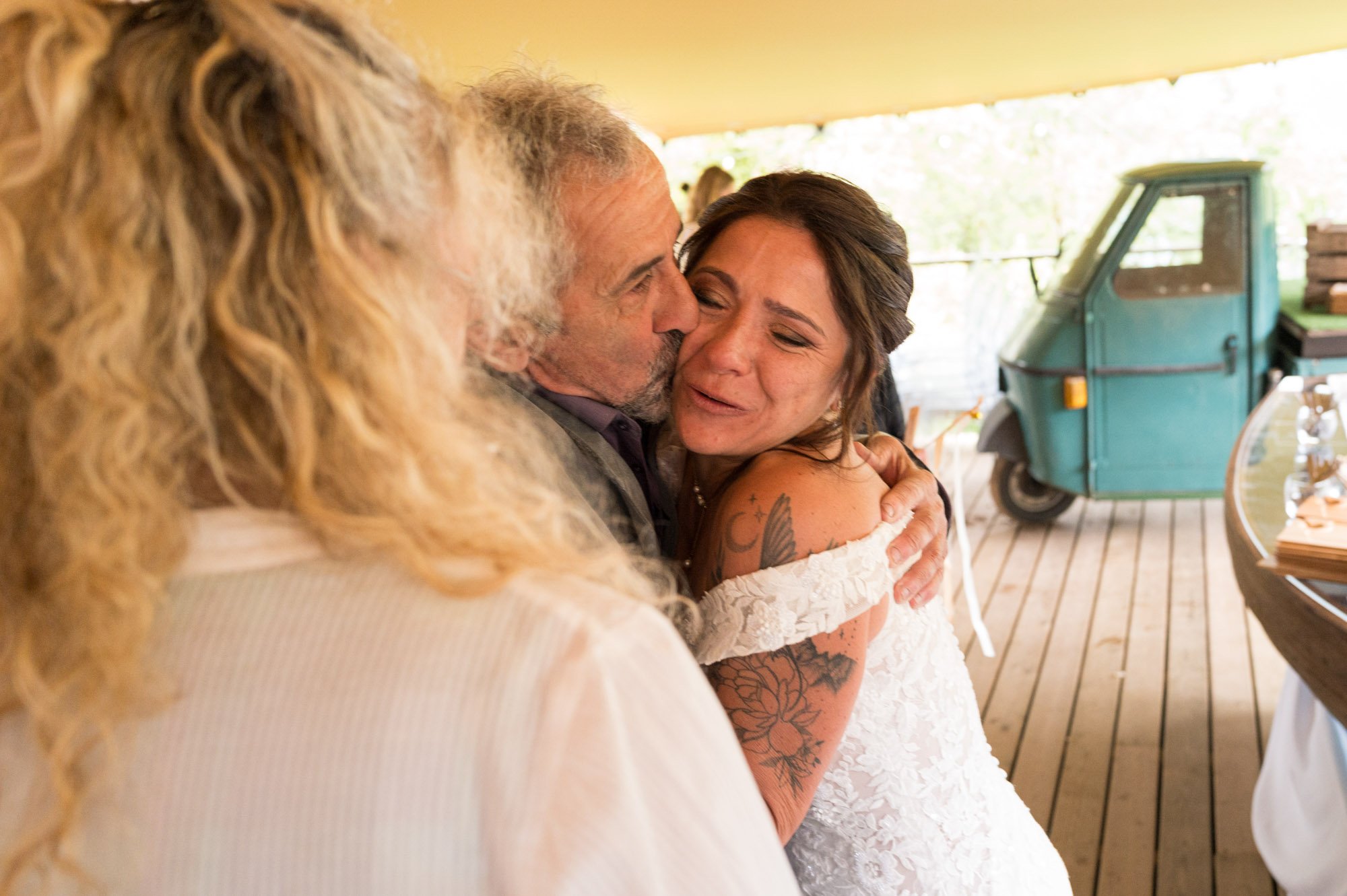 Un homme et une femme se font un câlin et un baiser lors d'une célébration de mariage, avec une autre femme aux cheveux bouclés en arrière-plan, dans un lieu avec un ponton en bois et un véhicule peu visible en arrière-plan.