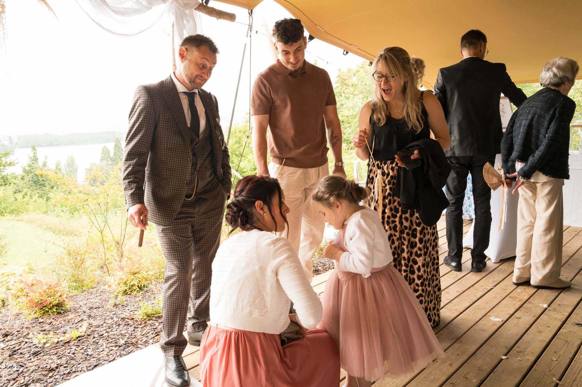 Groupe de personnes lors d'une fête ou célébration en plein air, sous une tente, avec vue sur la nature en arrière-plan, certaines discutant, d'autres regardant quelque chose à terre.