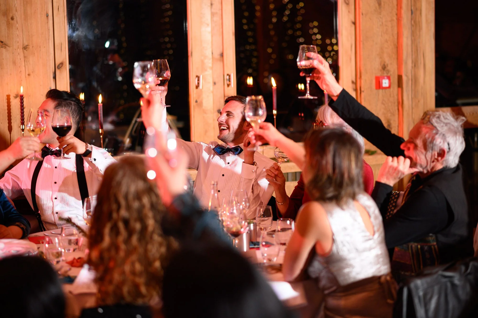 Groupe de personnes à un dîner festif, levant leurs verres de vin pour porter un toast, ambiance chaleureuse avec bougies et décoration en bois.