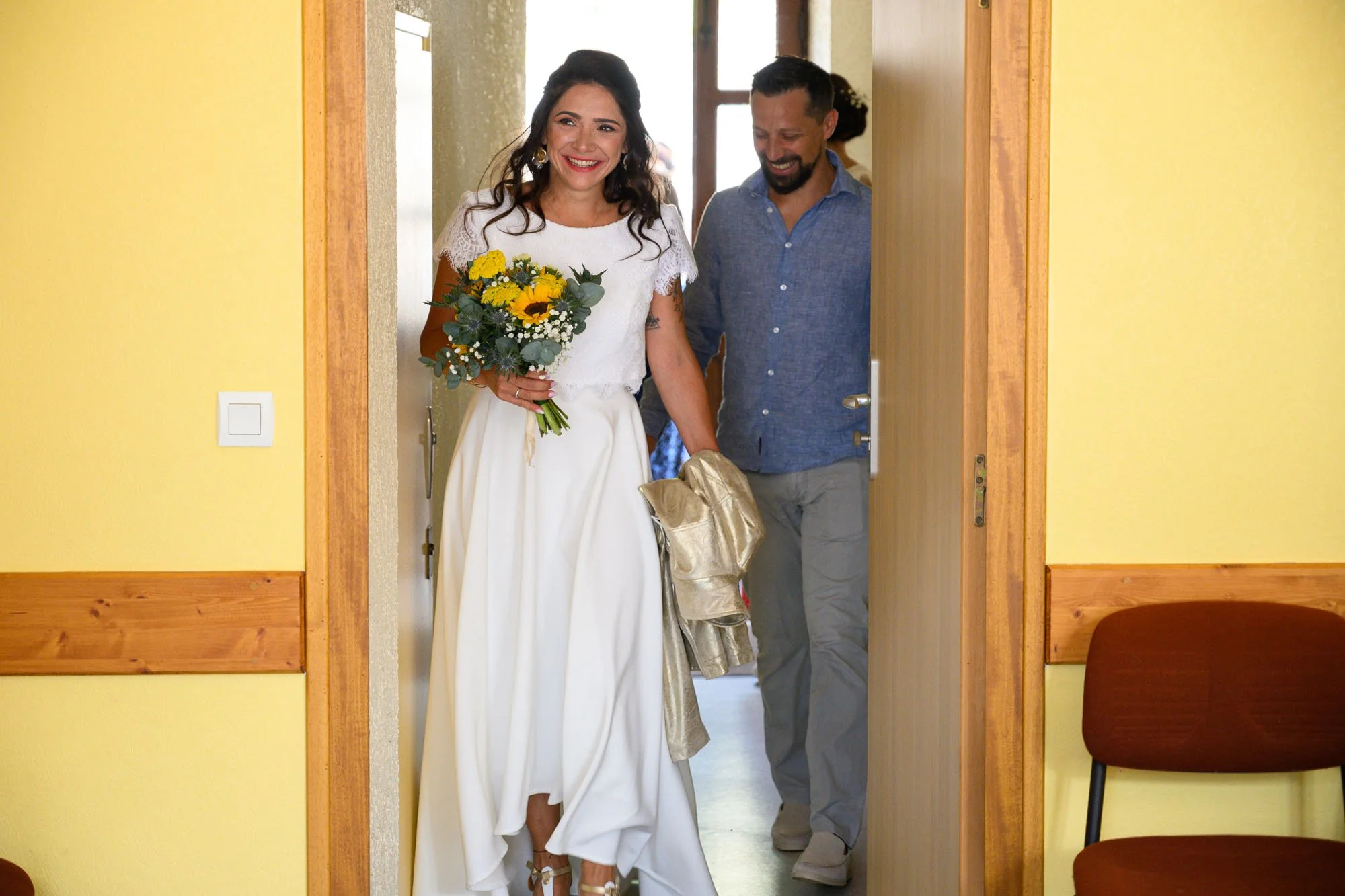 Une femme en robe blanche tenant un bouquet de tournesols et d'autres fleurs, joyeuse, entre dans une pièce, accompagnée d'un homme derrière elle.