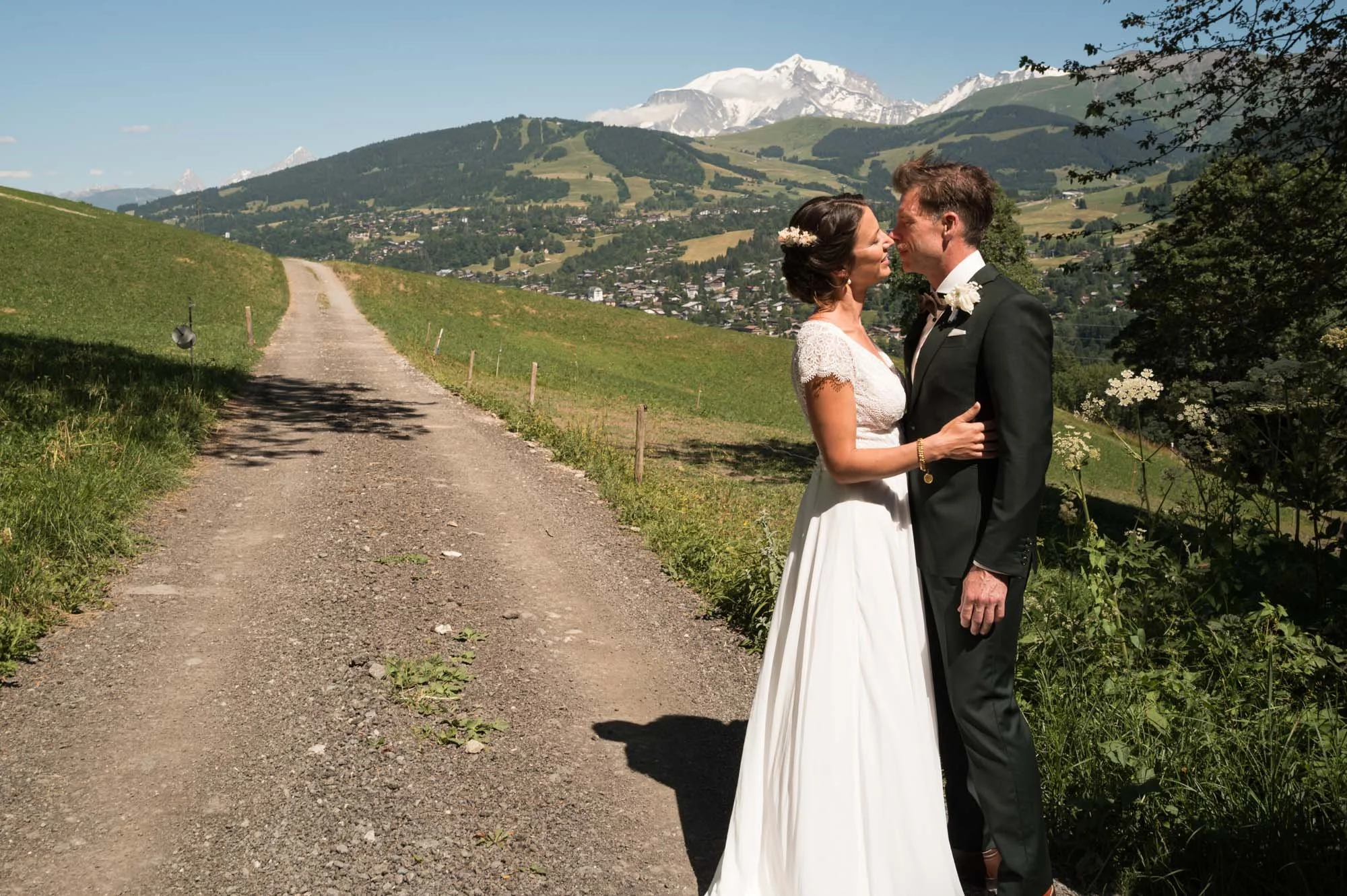 Un couple en vêtements de mariage, la femme en robe blanche et l'homme en costume noir, se tiennent près d'un chemin de terre dans un paysage de campagne verte avec des montagnes enneigées au fond.