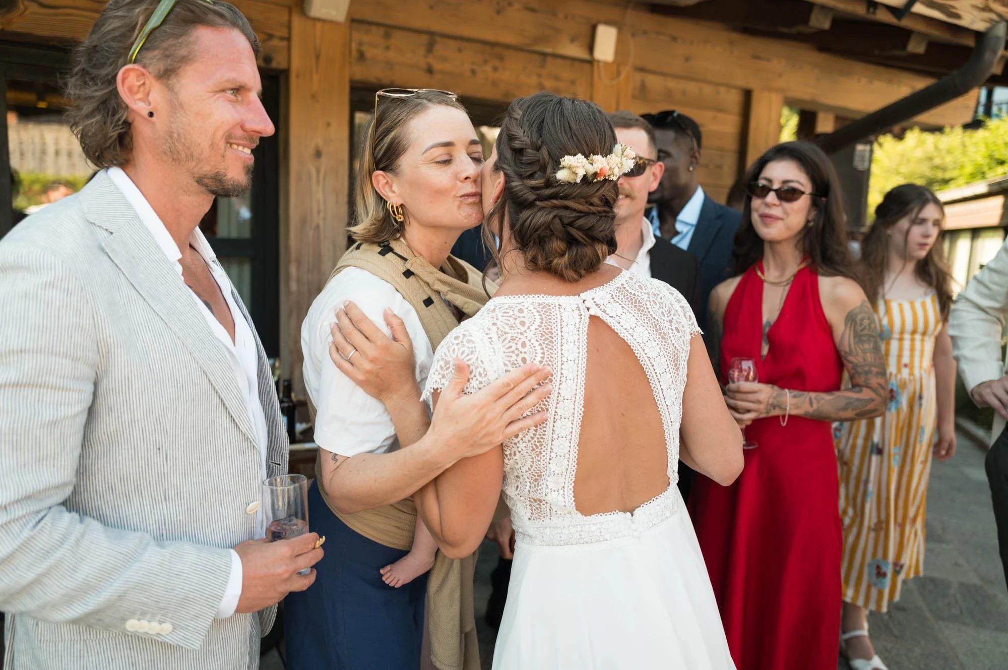 Un groupe de personnes en tenues de fête, dont une femme en robe de mariée embrassant une autre femme, lors d'un mariage en extérieur.