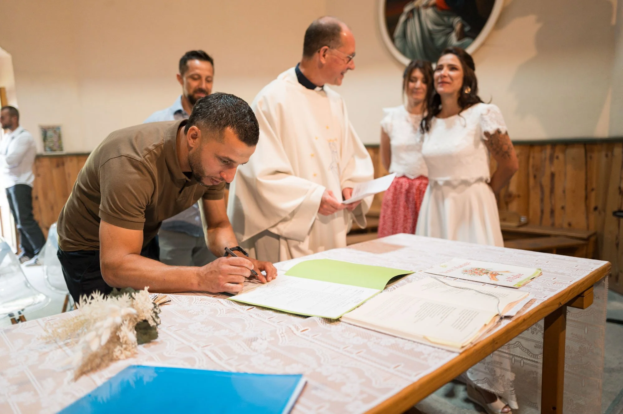 Un homme signe un document sur une table lors d'une cérémonie dans une salle décorée en bois, entouré de personnes en vêtements formels et religieux, avec un miroir rond en arrière-plan.