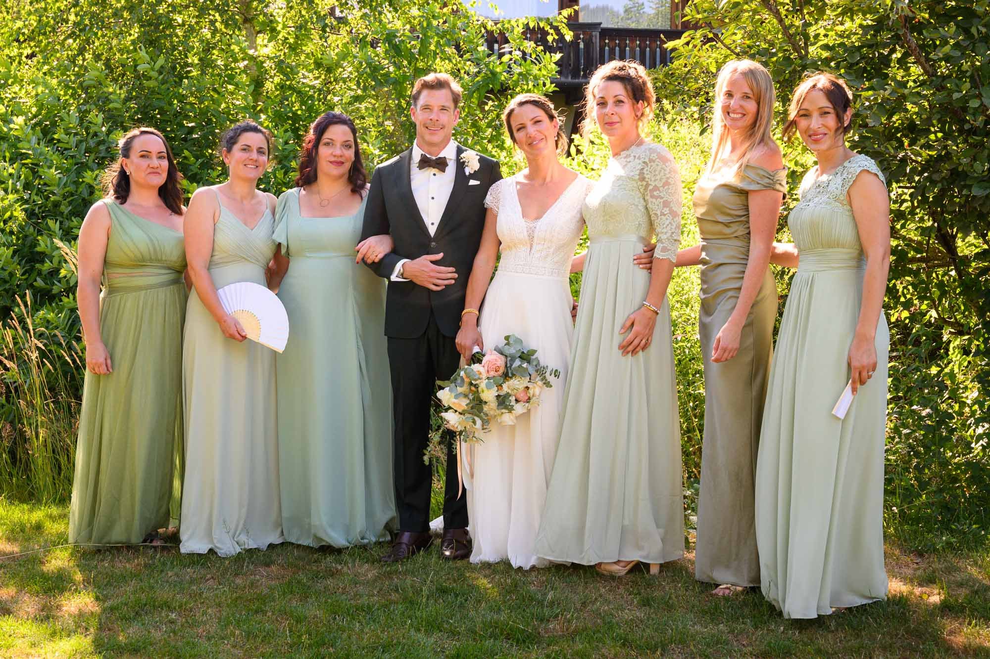 Group de personnes en tenue de mariage posant dans un jardin ensoleillé, avec vegetation verte en arrière-plan.