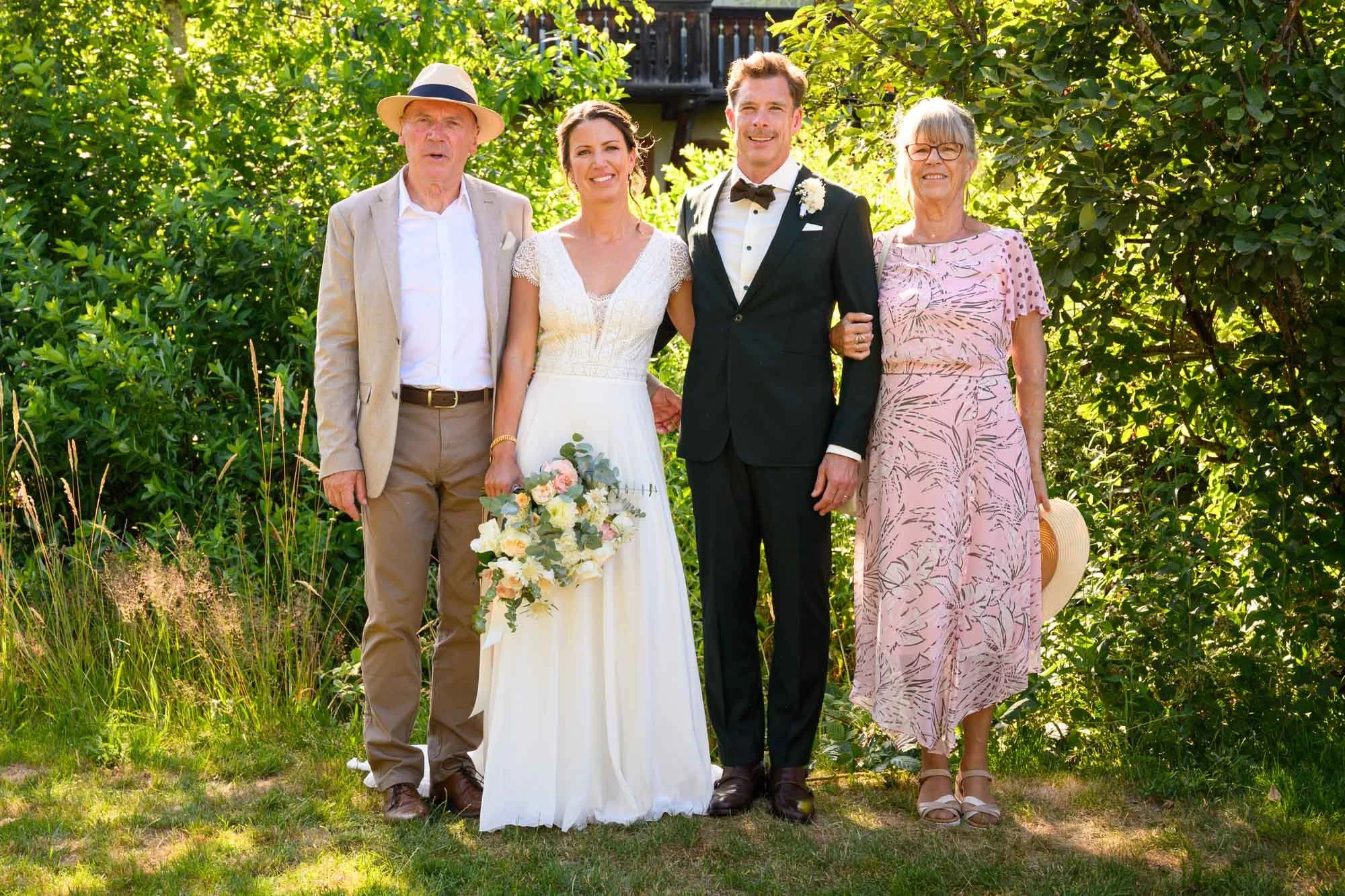 Groupes de cinq personnes posant pour une photo lors d'un mariage en plein air, avec deux femmes âgées et un homme âgé, un couple de jeunes mariés, et un arrière-plan de verdure.
