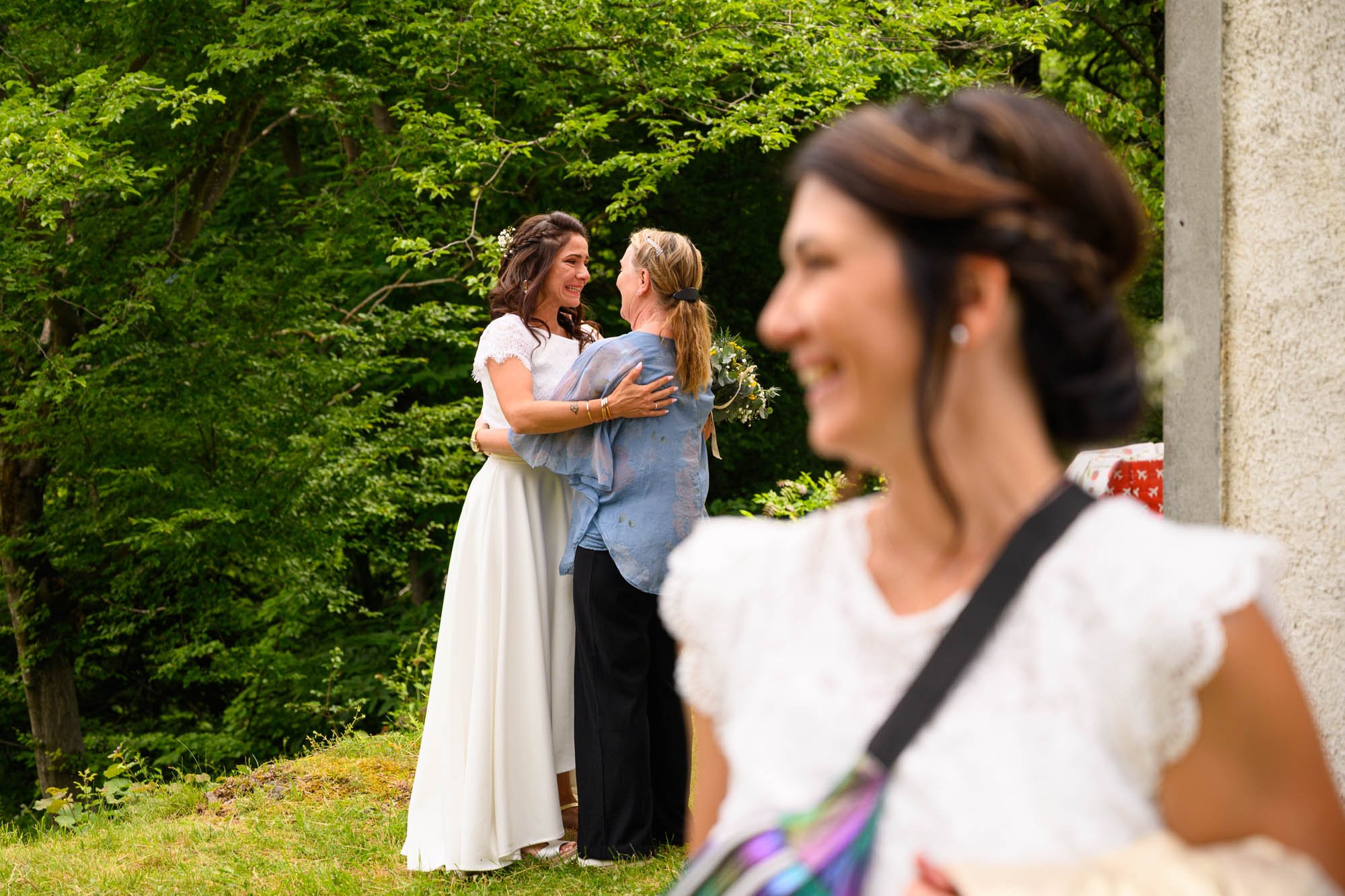 Une femme en robe de mariage heureuse embrassant une autre femme lors d'une cérémonie en plein air, avec une femme en premier plan floue qui sourit.