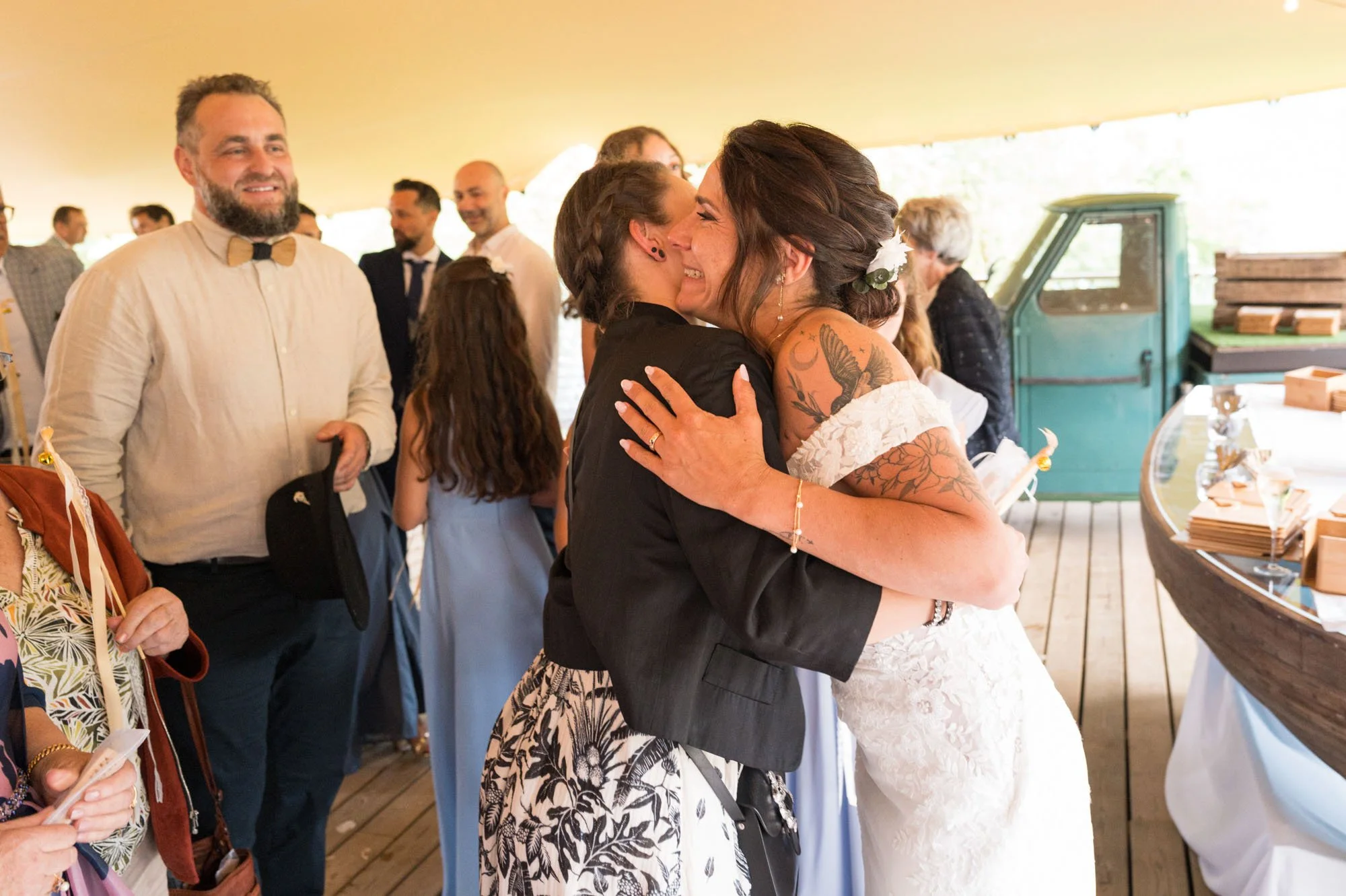 Deux femmes en robe de mariage et en costume s'embrassent et se font un câlin lors d'une célébration nuptiale, entourées d'invités lors d'une fête en intérieur.