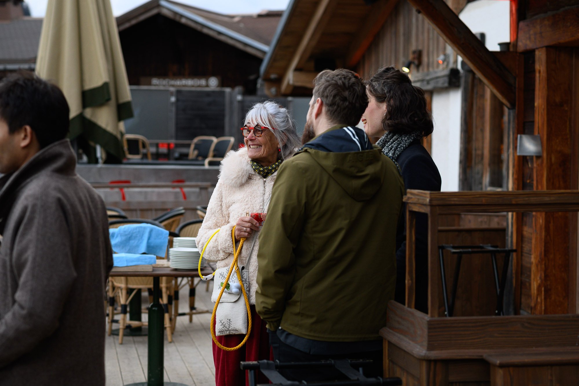 Groupe de personnes discutant et souriant en extérieur devant des bâtiments en bois, vêtements chauds, ambiance conviviale.