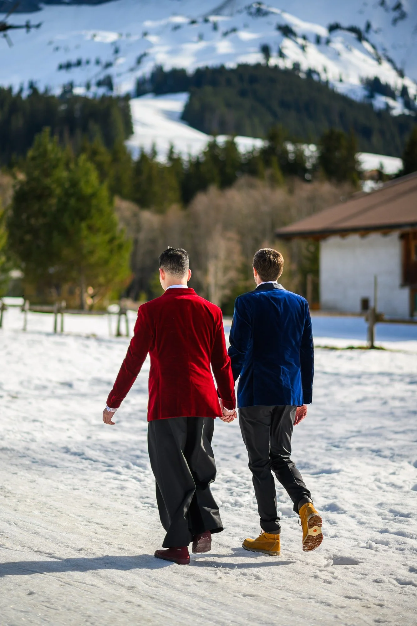 Deux hommes en manteaux rouges et bleus marchant dans la neige, montagnes enneigées en arrière-plan.