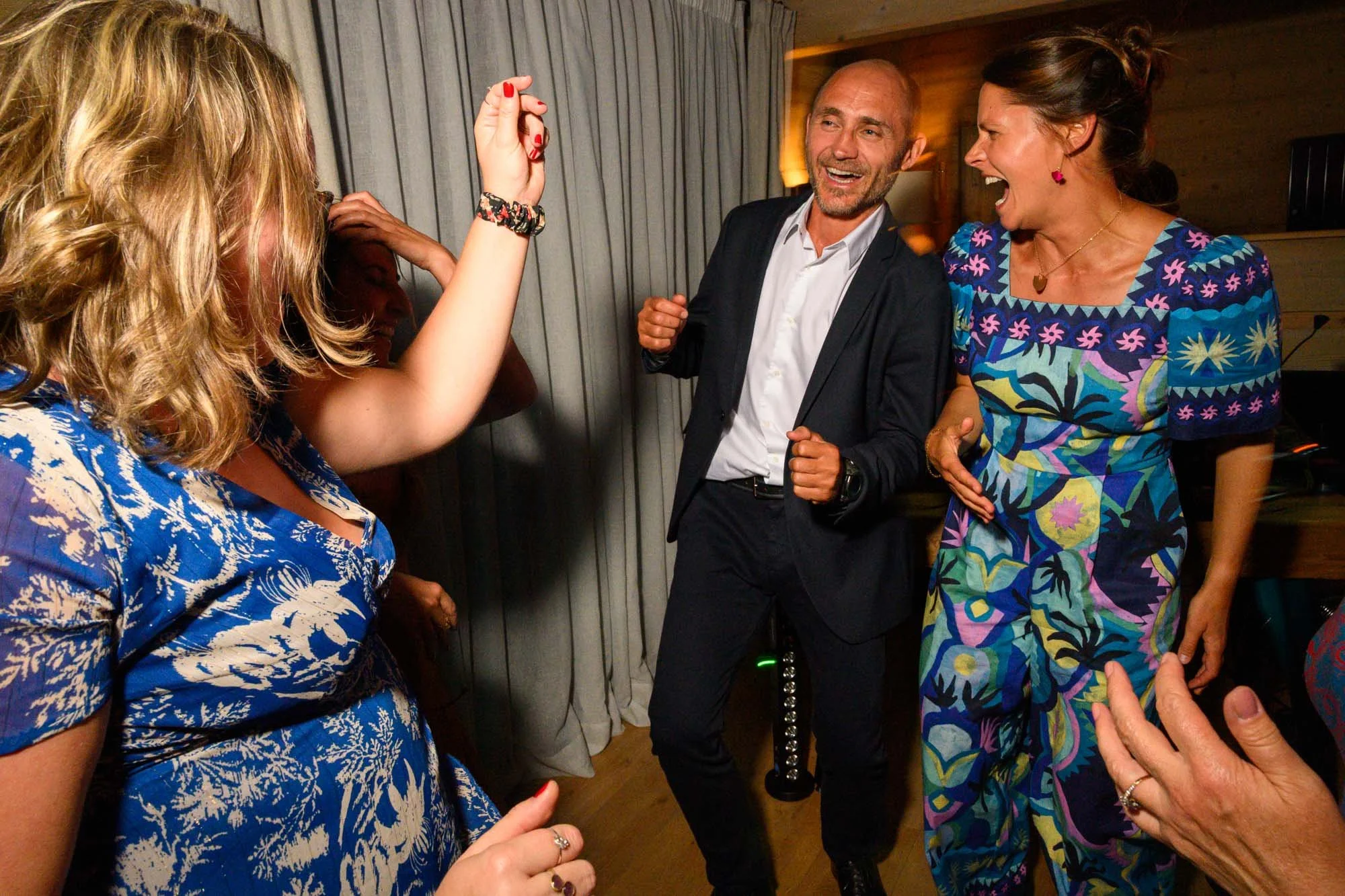 Quatre personnes souriantes et riantes dans une fête ou un rassemblement, dans une pièce avec un mur en bois et des rideaux gris, en train de danser ou de s'amuser ensemble.