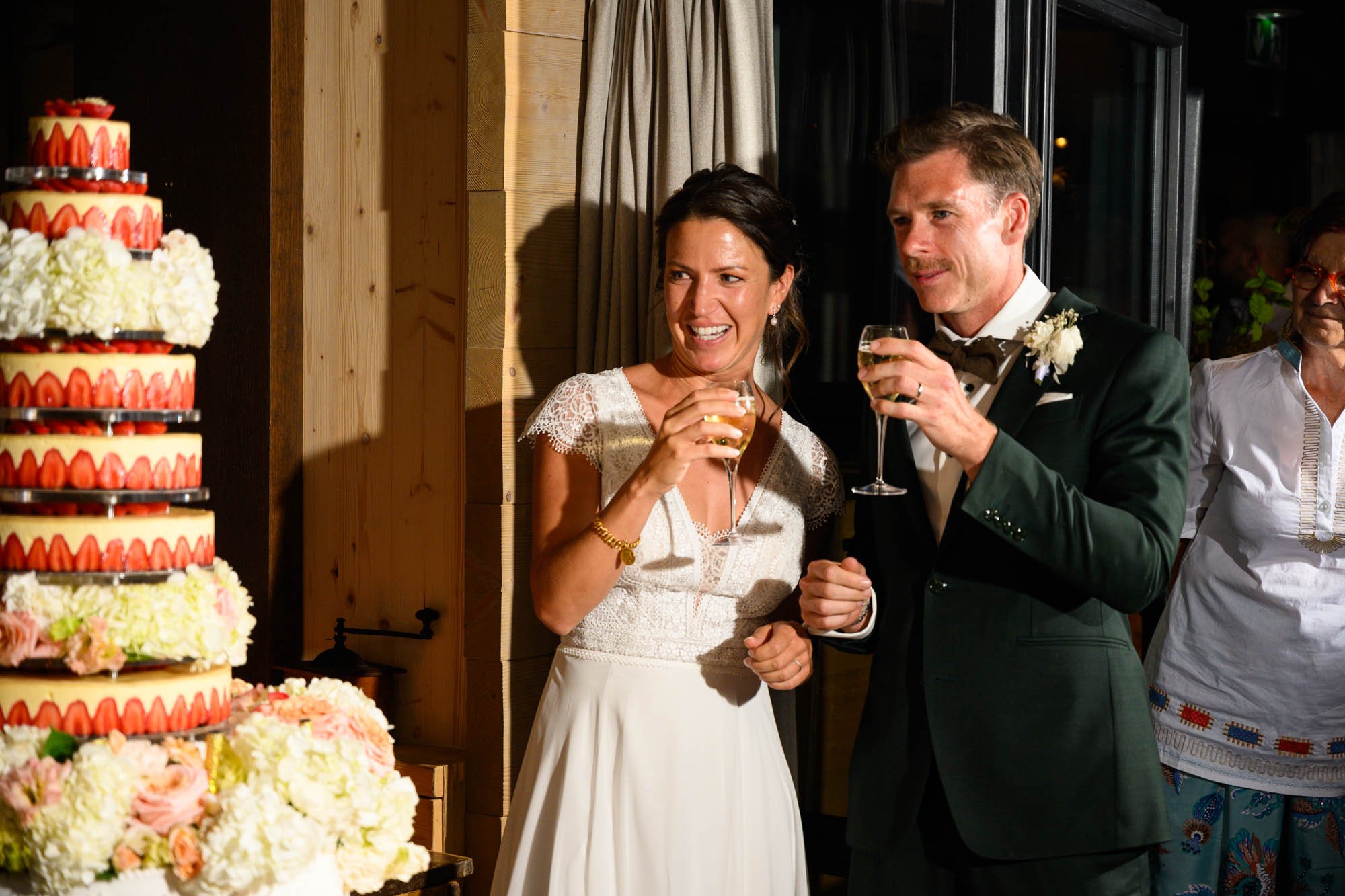 Un couple de mariés souriants, tenant des verres de champagne, se célèbre lors d'un mariage avec un gâteau à plusieurs étages décoré de fraises et de fleurs, à l'intérieur d'une salle de réception en bois.
