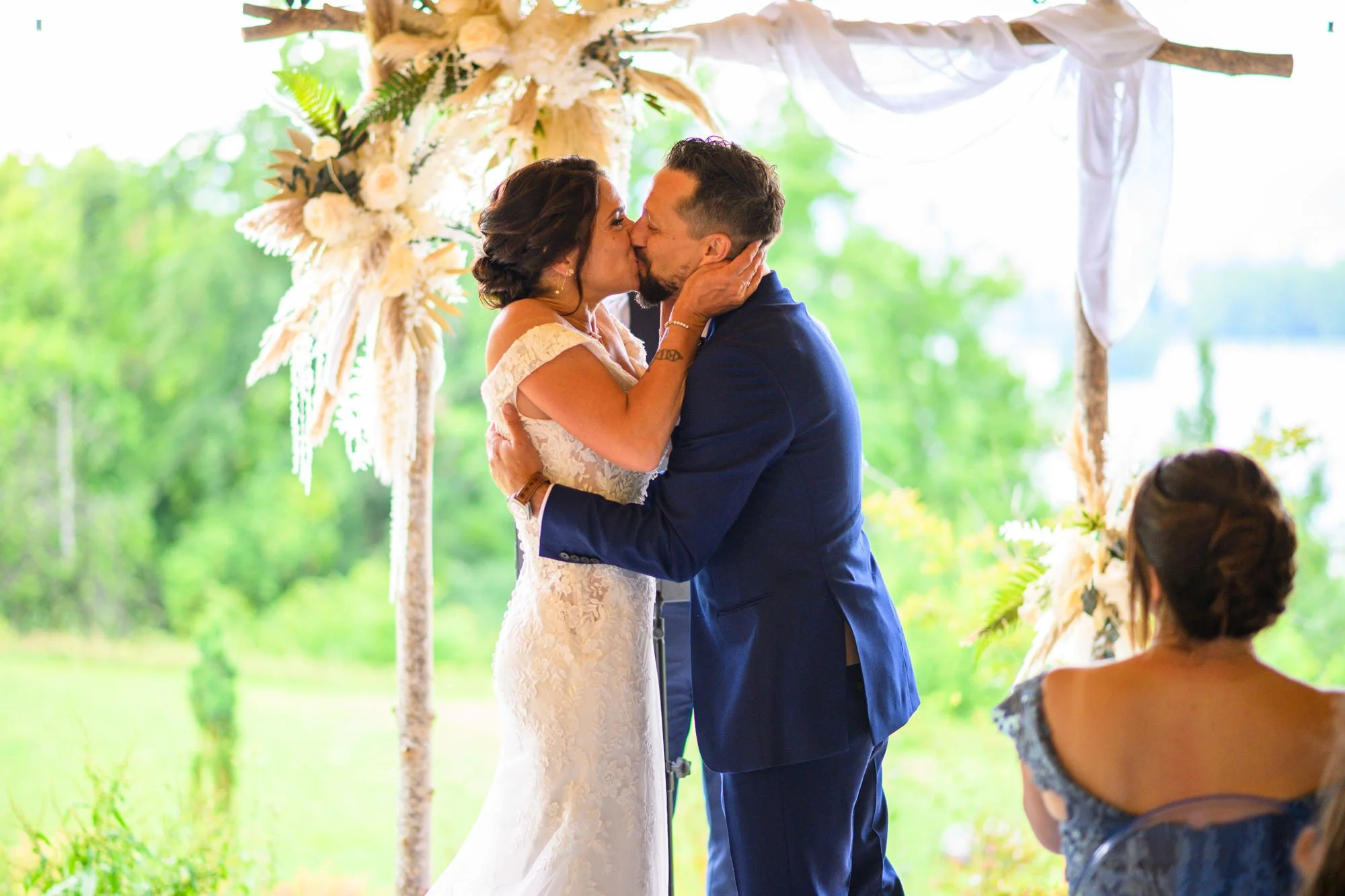 Un couple en mariage s'embrassent lors de la cérémonie à l'extérieur entouré de verdure, avec une femme assise regardant la scène.