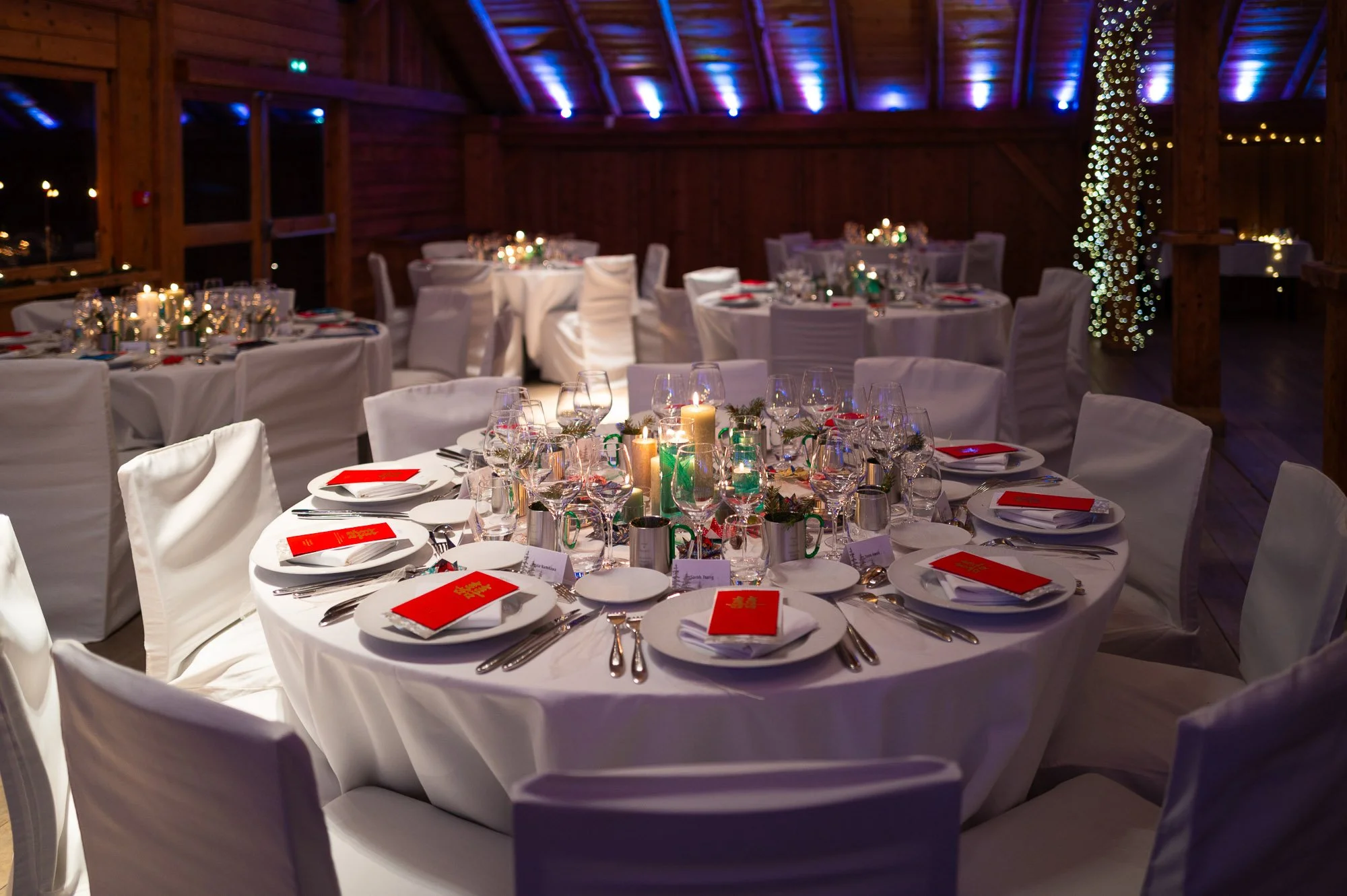 Salle de réception décorée pour un dîner, avec tables rondes habillées en blanc, nappes blanches, verres à vin, assiettes, couverts, bougies et petits centres de table, ambiance chaleureuse avec éclairage tamisé et guirlandes lumineuses.