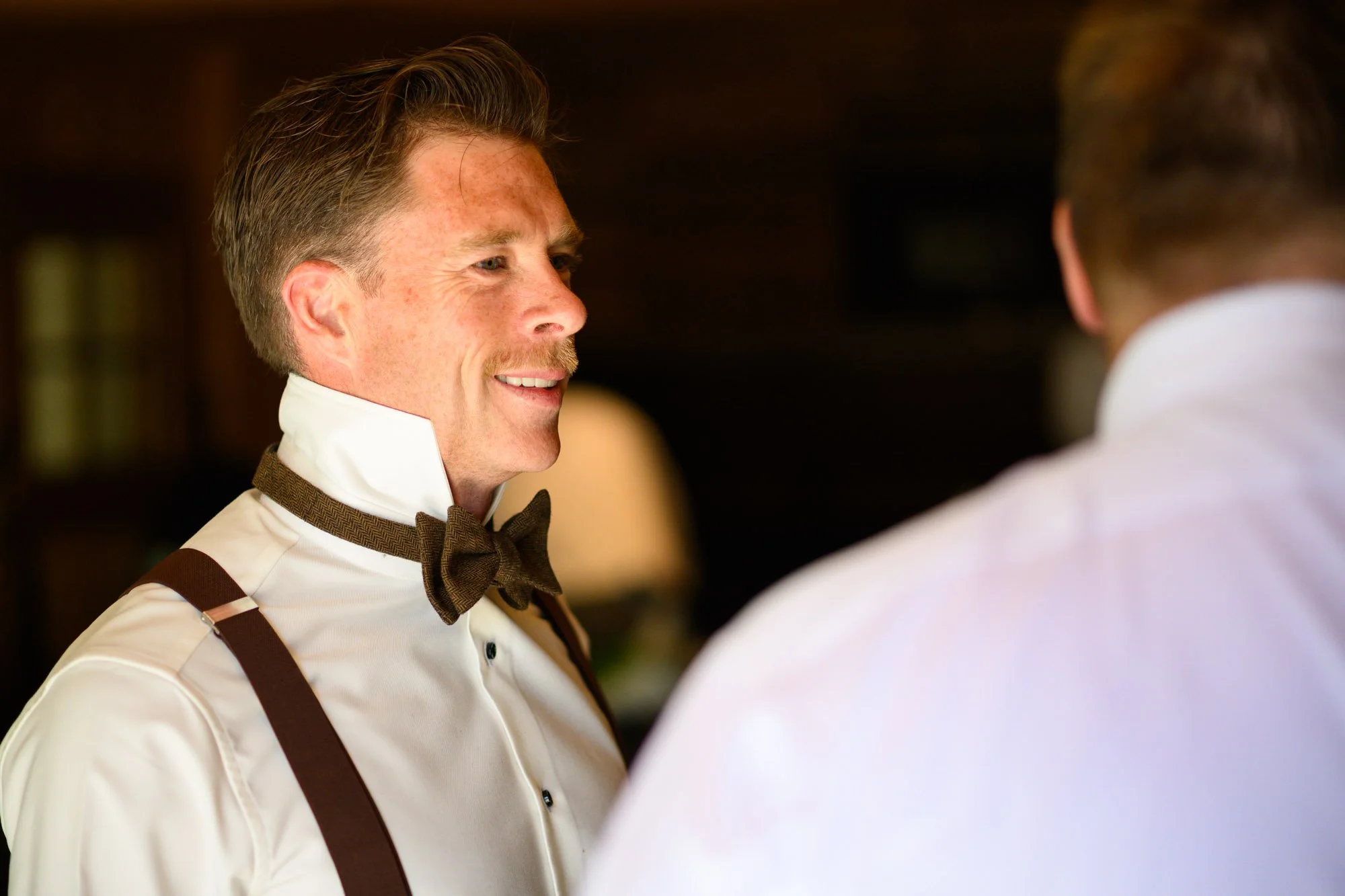 Un homme souriant, habillé en chemise blanche avec une lavallière marron, discutant avec une autre personne dont le dos est visible.