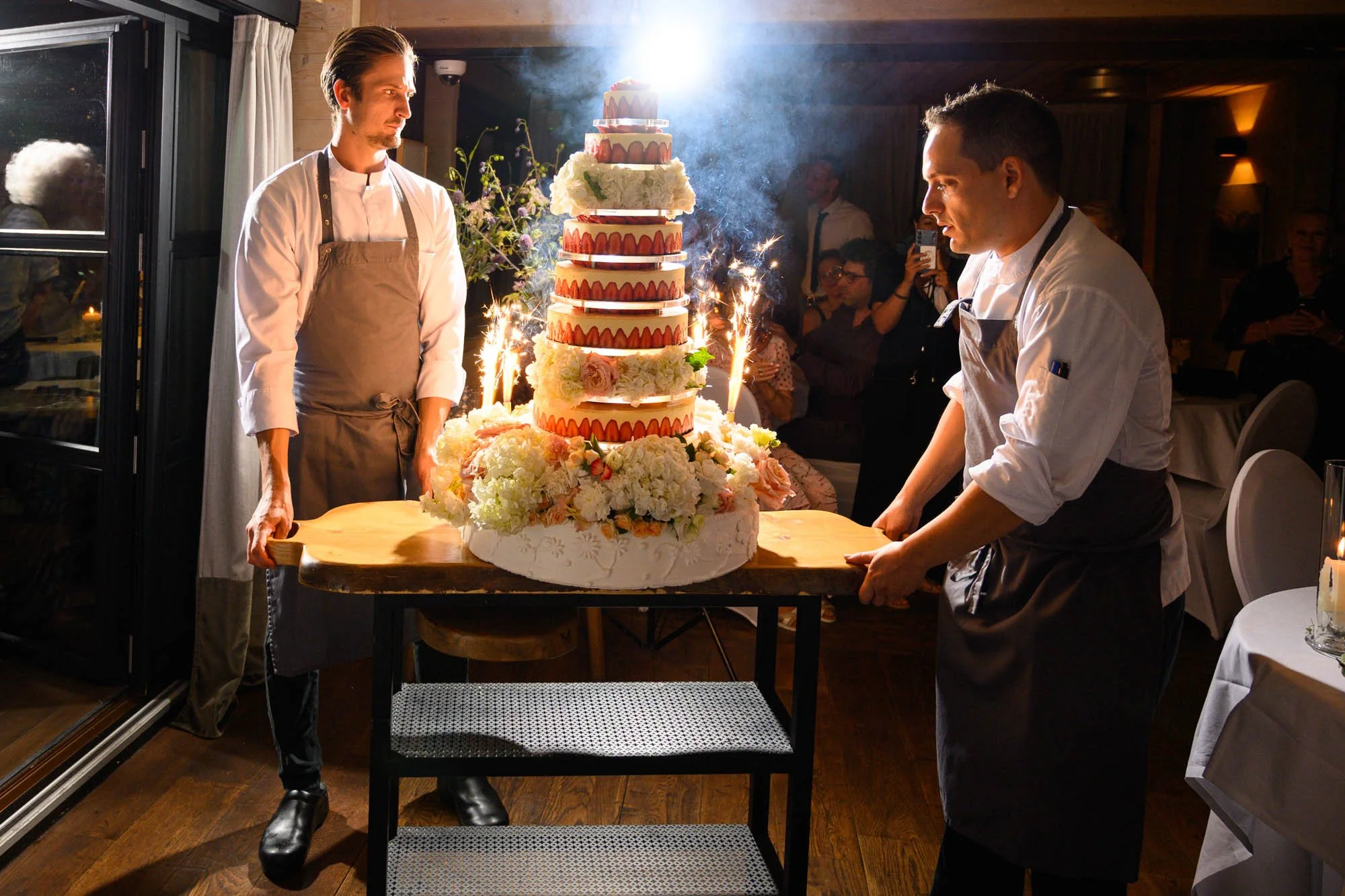 Deux chefs manipulent une grande pièce montée de gâteau de mariage décorée de fleurs, avec des feux d'artifice et de la fumée, lors d'une célébration dans une salle avec des invités en arrière-plan.