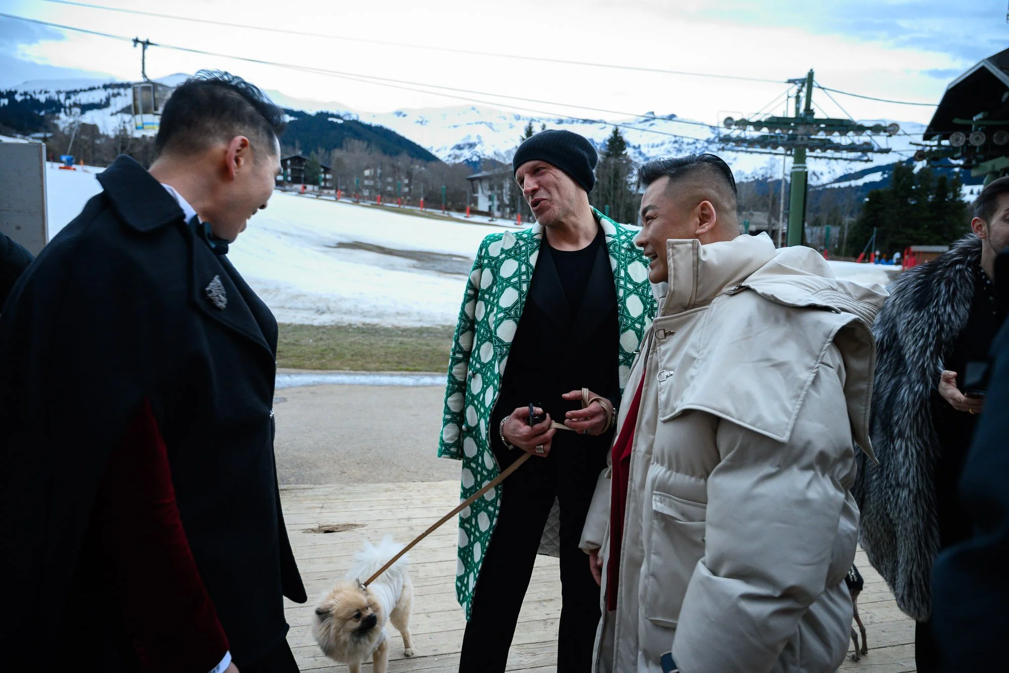 Groupe de personnes en vestes d'hiver discutant près d'un chien à la station de ski, avec des montagnes enneigées en arrière-plan.