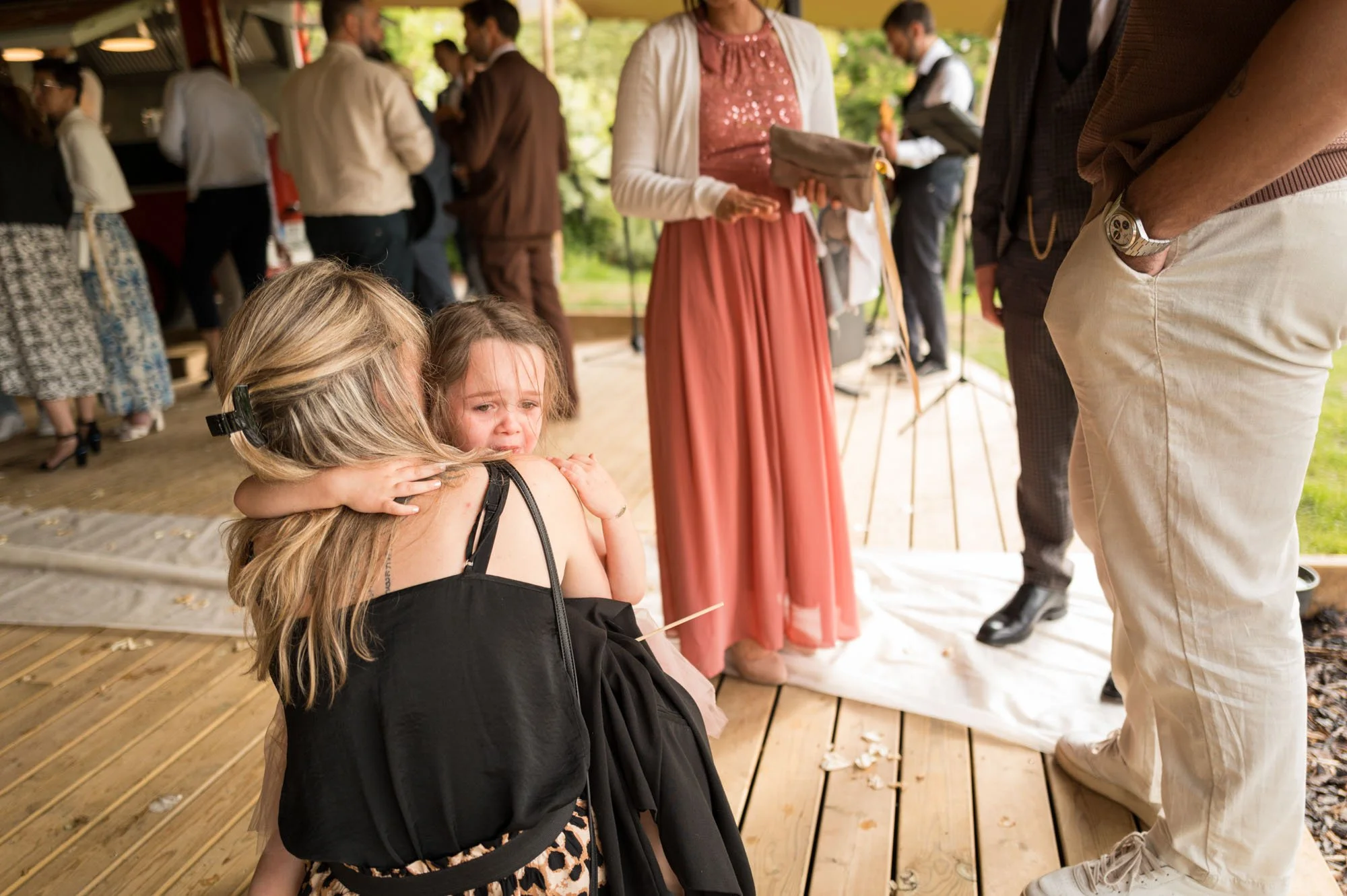 Une femme et une petite fille qui pleure, entourées de personnes lors d'une cérémonie en plein air, avec des invités en arrière-plan.