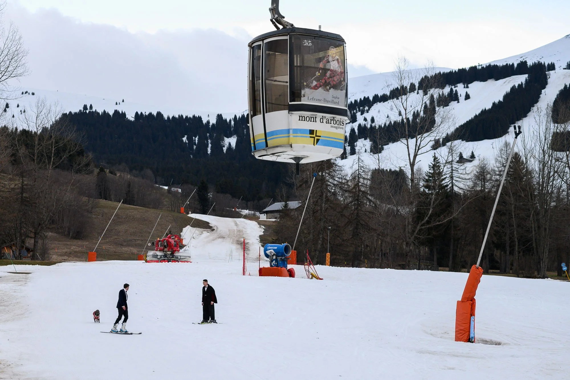 Deux personnes skiant sur une pente enneigée dans une station de ski, avec un téléphérique suspendu dans le ciel. La scène est entourée d'arbres et de montagnes enneigées.