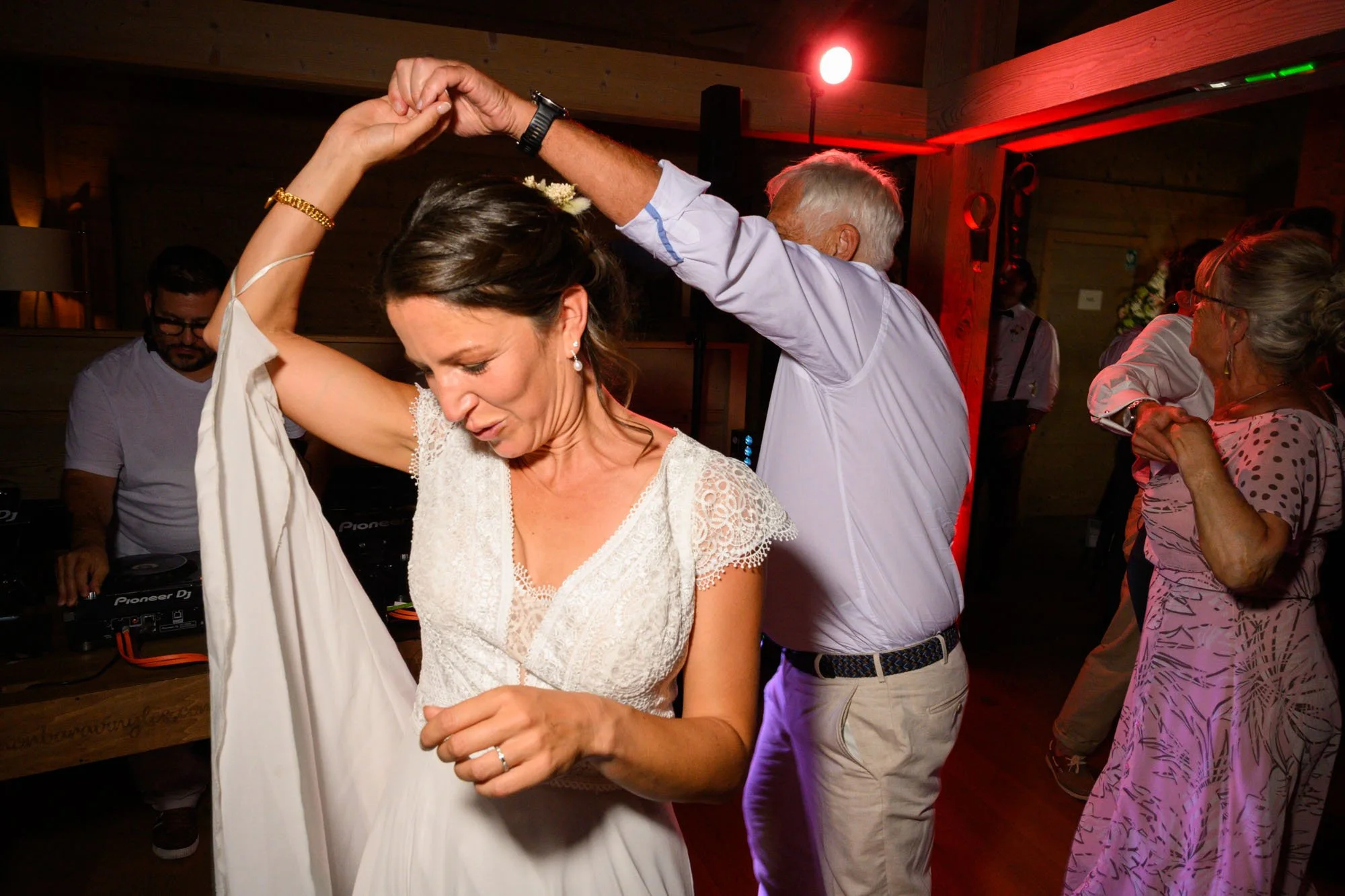 Une femme en robe blanche danse avec un homme lors d'une fête. D'autres personnes dansent en arrière-plan, ambiance chaleureuse avec éclairage tamisé et décor en bois.