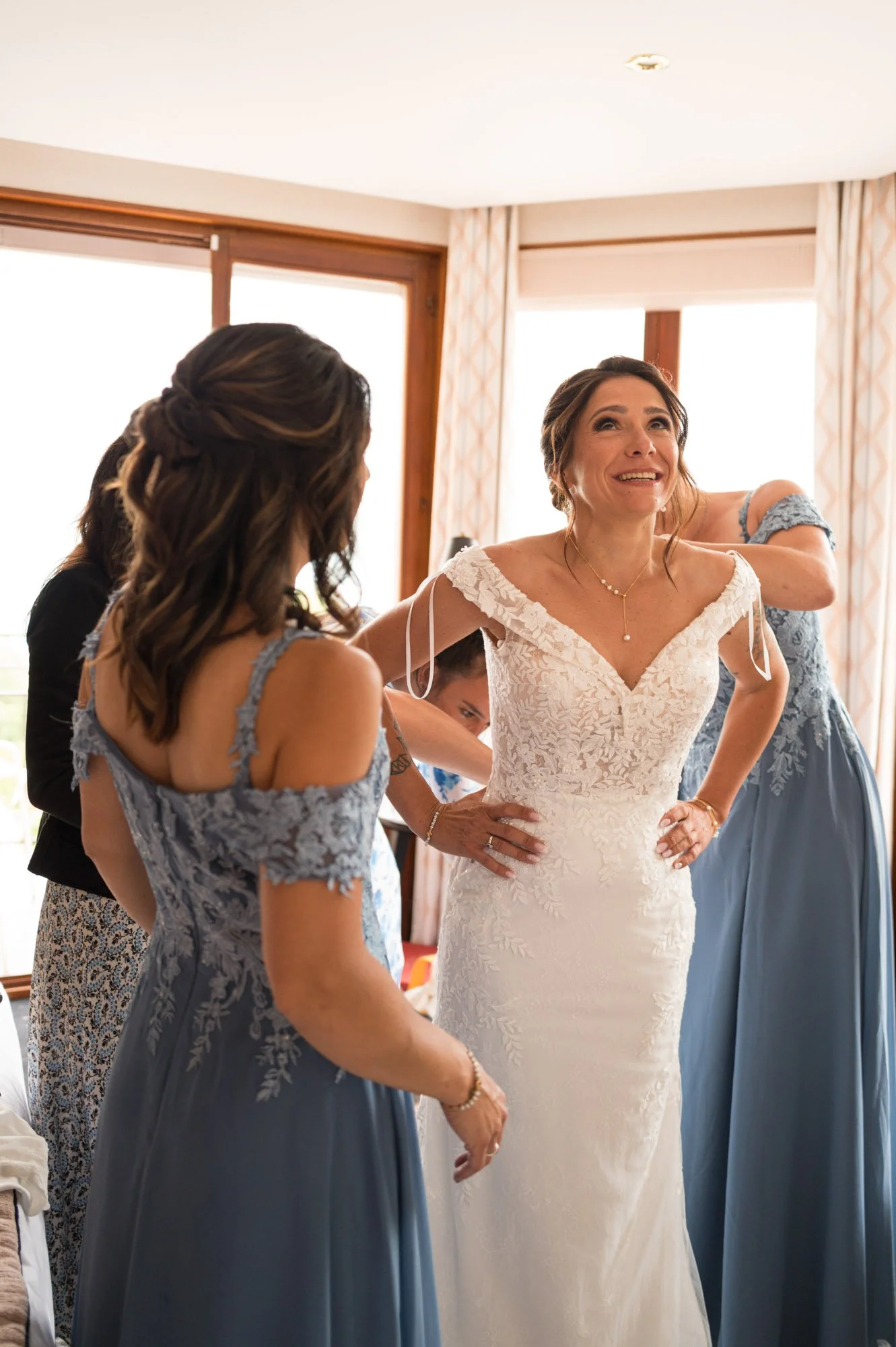 Une femme en robe de mariée sourit alors qu'elle est entourée de femmes qui l'aident à se préparer pour un mariage dans une pièce lumineuse avec des fenêtres.