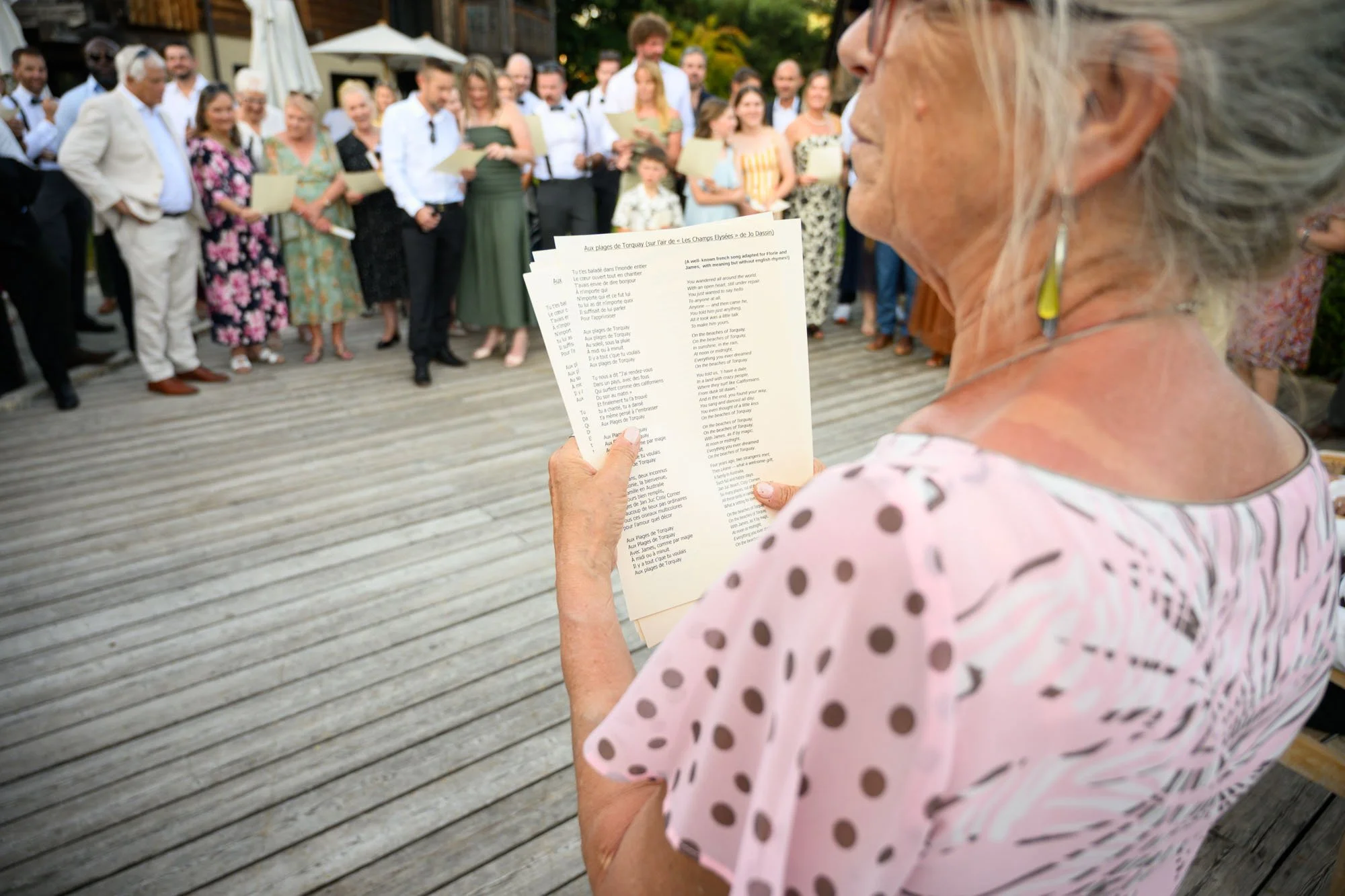Une femme âgée lit à haute voix des paroles de chanson lors d'une cérémonie ou d'une réunion en plein air, avec un groupe de personnes en tenue de fête en arrière-plan.