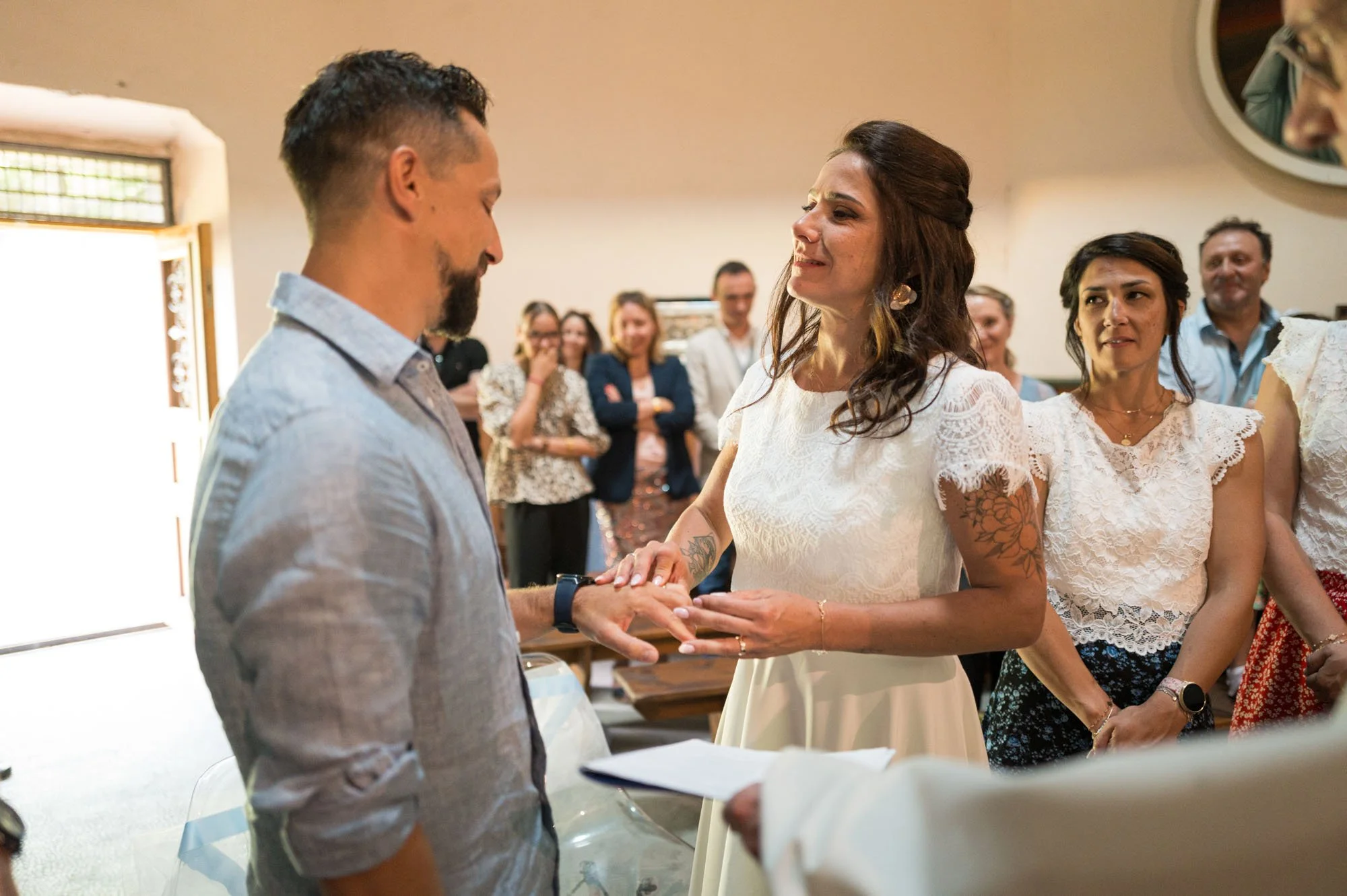 Une femme en robe blanche tenant la main d'un homme lors d'une cérémonie dans une pièce avec plusieurs personnes en arrière-plan