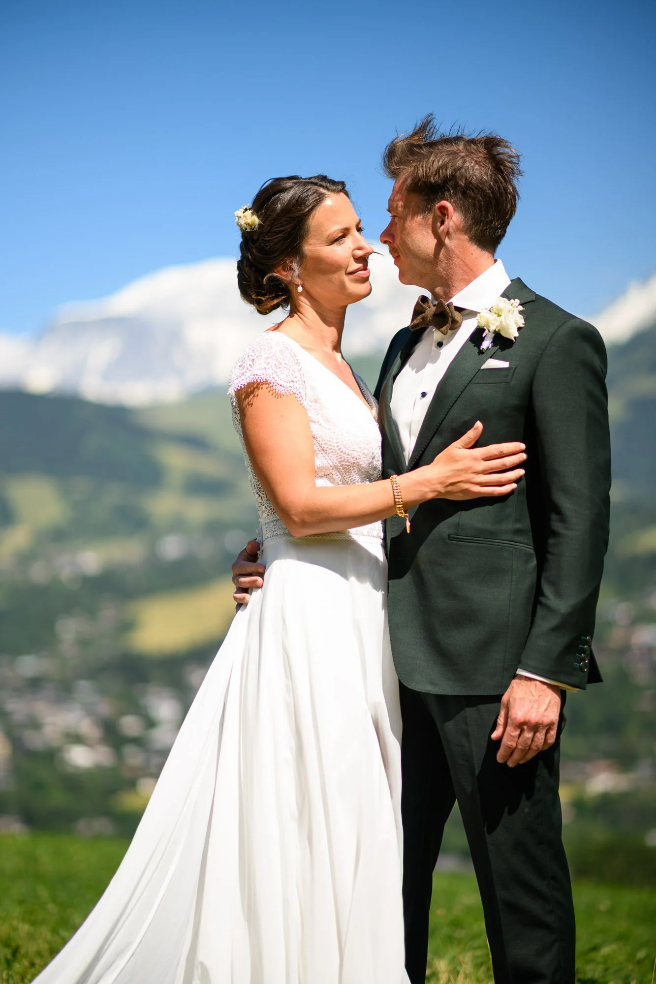 Un couple en costume de mariage, une femme en robe blanche et un homme en smoking, se regardant tendrement contre un paysage de montagnes et un ciel bleu.
