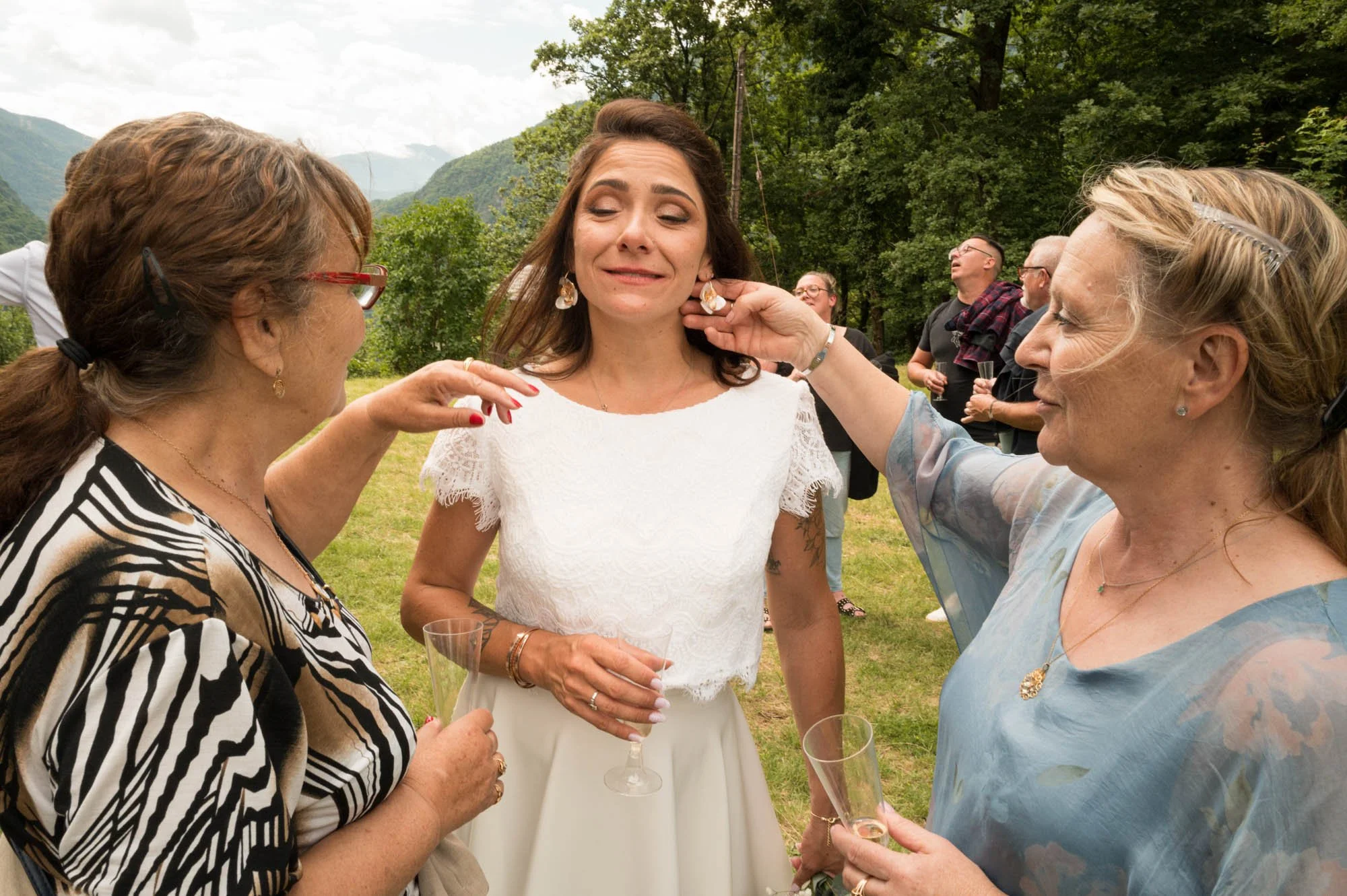 Une femme en robe blanche est entourée de deux autres femmes lors d'une fête en plein air, en train de recevoir des conseils ou des compliments, avec un groupe de personnes en arrière-plan et un paysage de montagnes et de verdure.