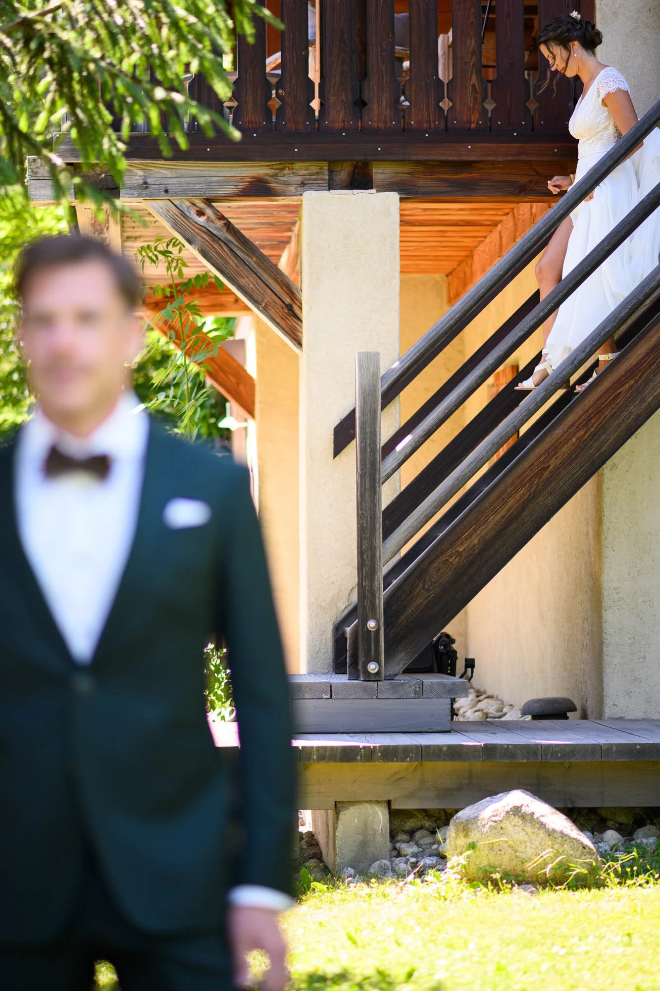 Une femme en robe blanche descend un escalier en bois, en arrière-plan, un homme en costume sombre et nœud papillon est flou devant un peu d'herbe verte.
