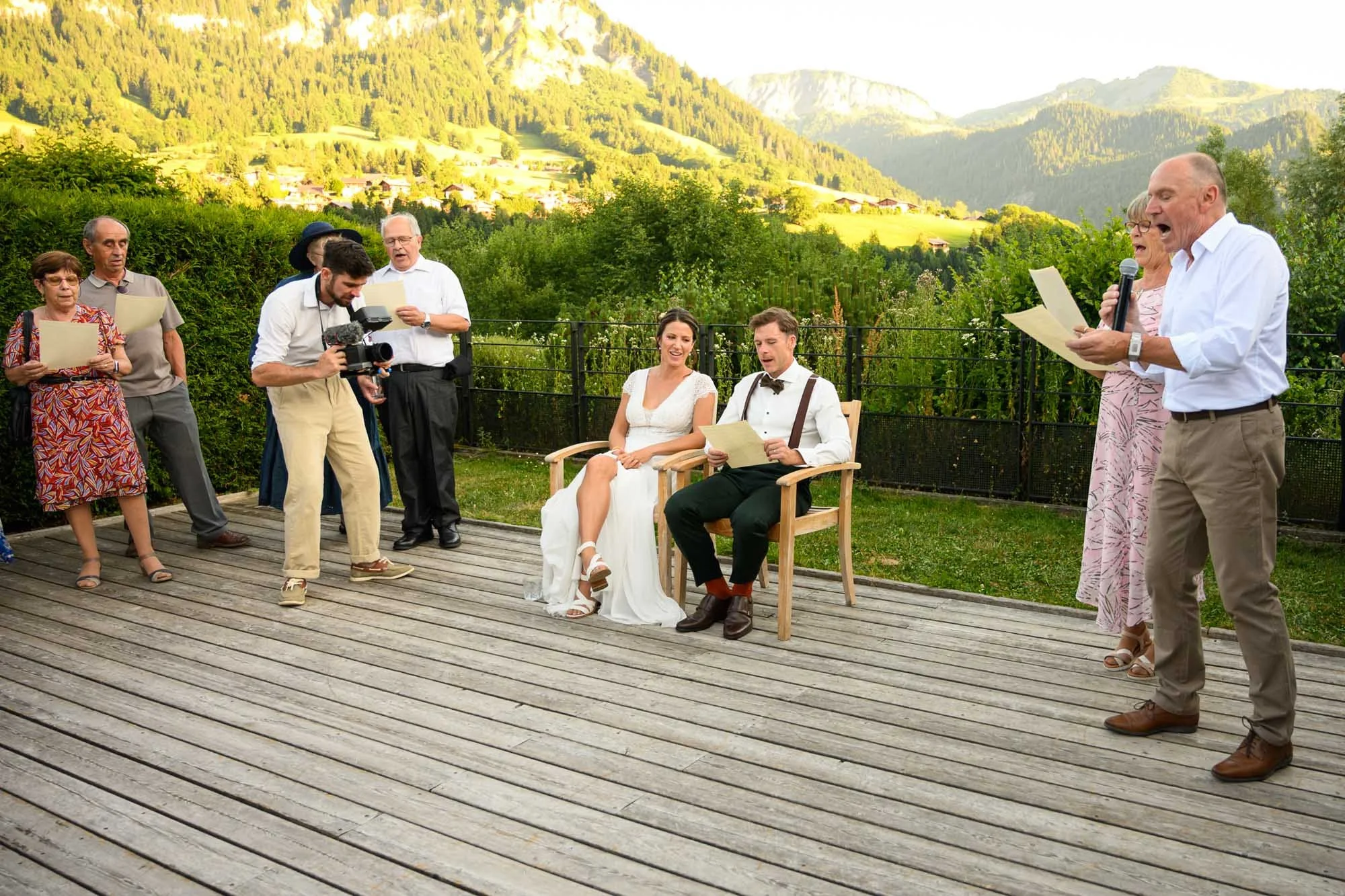 Cérémonie de mariage en plein air avec un couple assis, entouré de plusieurs personnes qui lisent ou enregistrent, sur un pont en bois avec un paysage de montagnes et de verdure en arrière-plan.